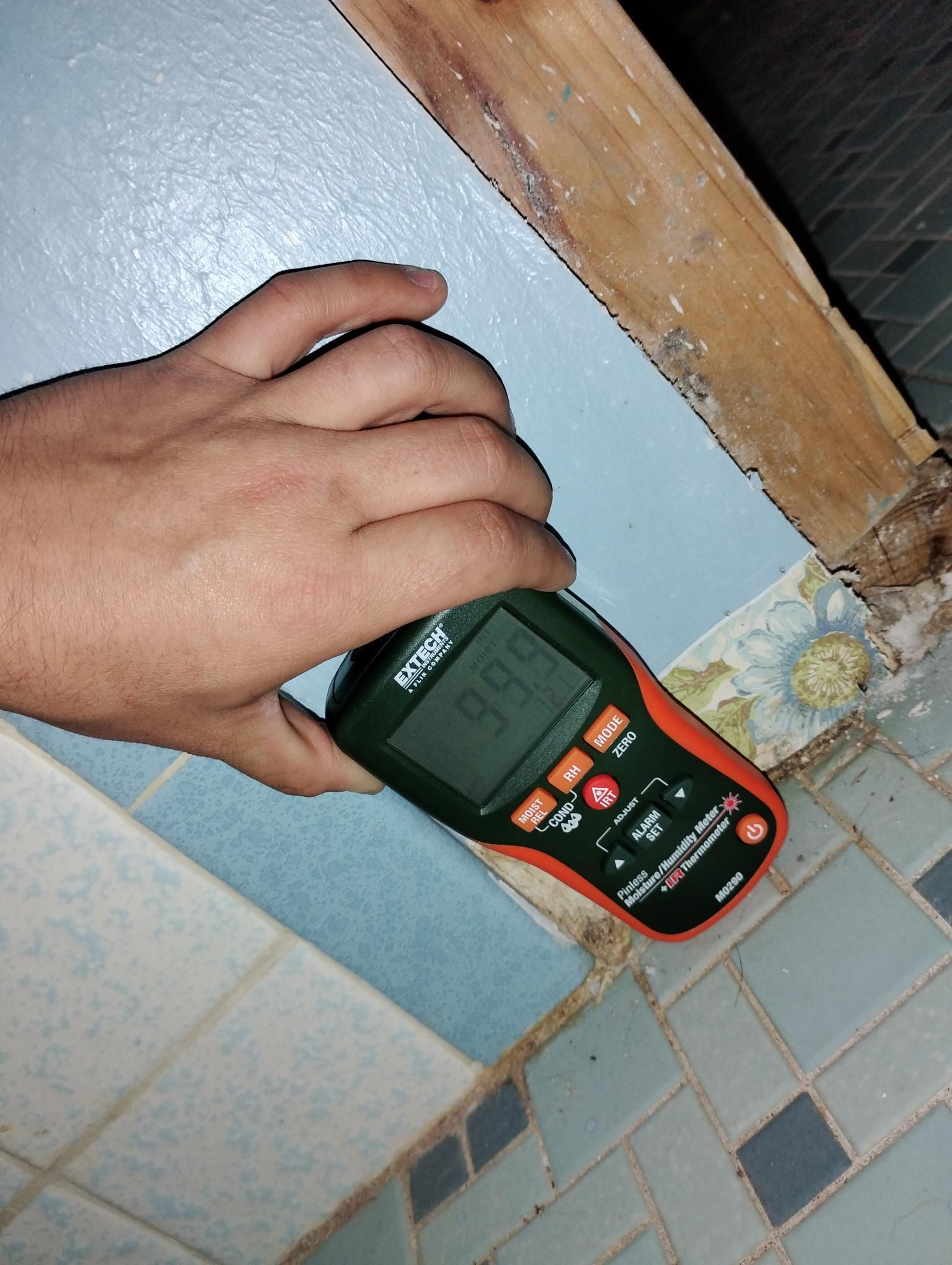Hand holding a moisture meter against a blue bathroom wall, reading 99.7%. Wooden frame and tile visible.