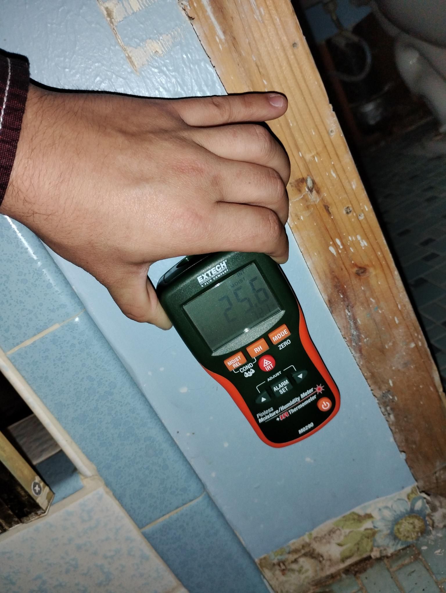 Hand holding a moisture meter on a blue-tiled surface and wooden frame, reading 25.6.