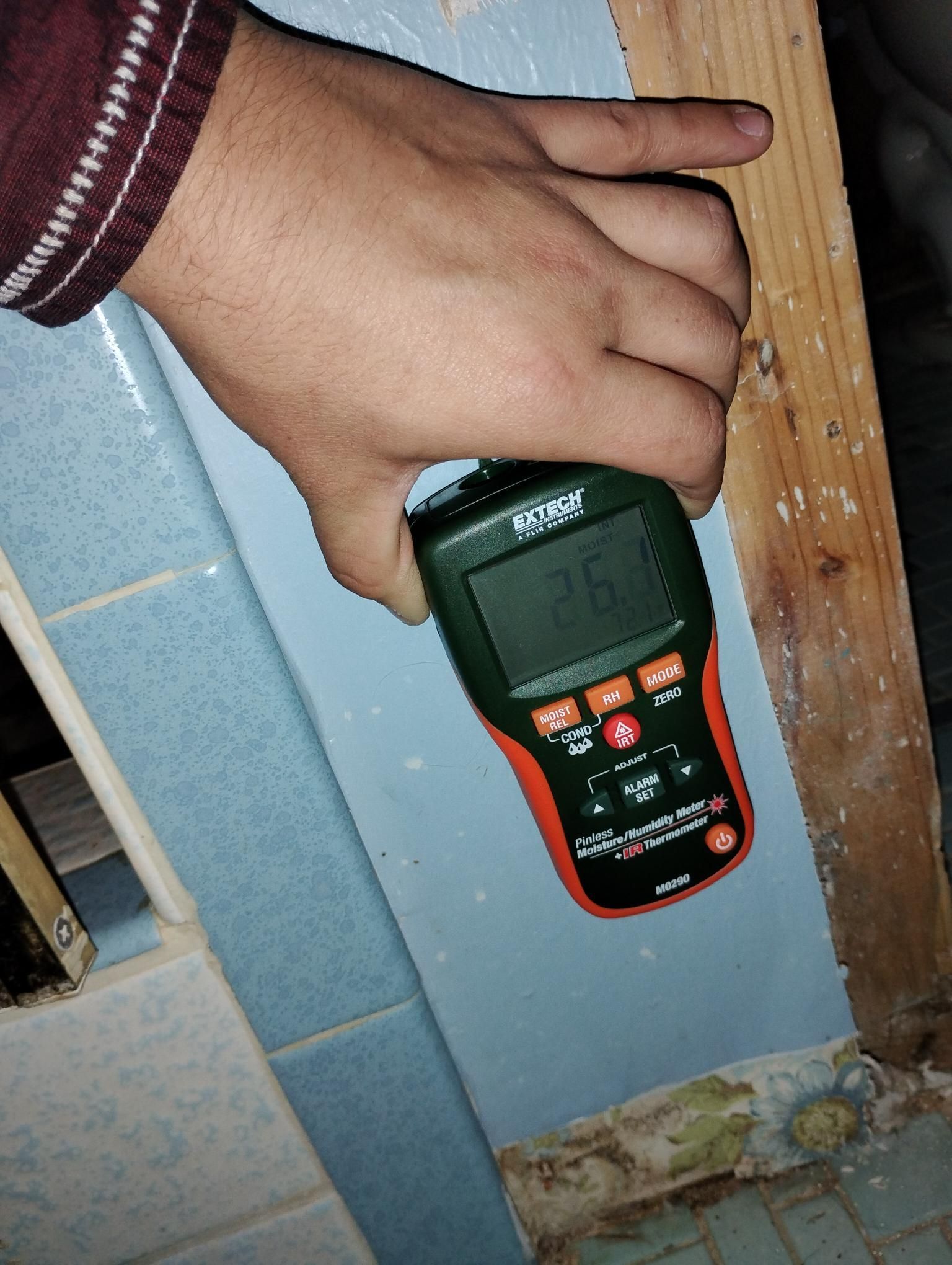 Hand holding a moisture meter against blue tile in a bathroom; meter reads 26.