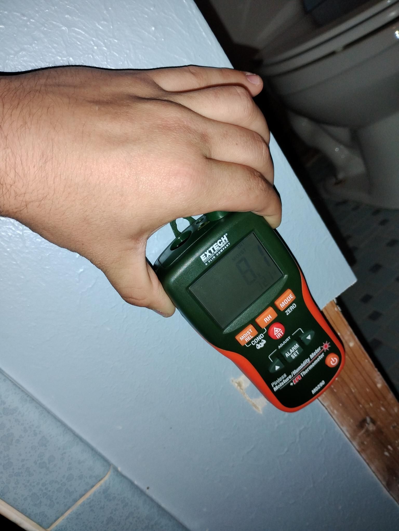 Hand holding a green moisture meter against a blue painted wall in a bathroom.