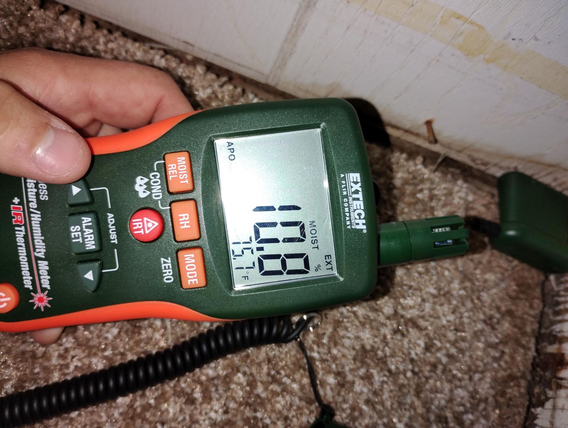 Hand holding a moisture meter on carpet near a wall, displaying a reading of 10.8.