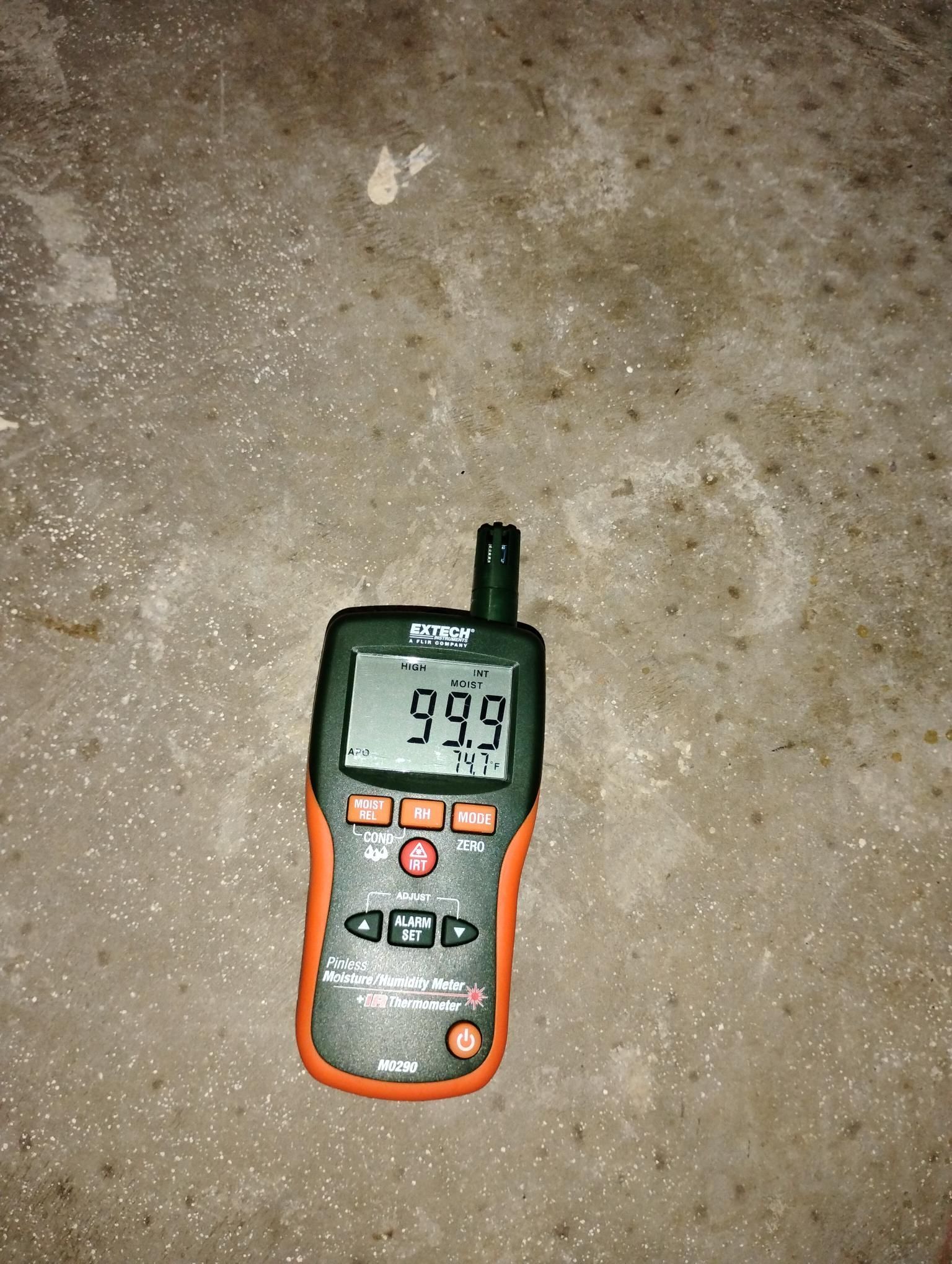 Orange and black humidity meter showing 99.9 on a mottled gray concrete surface.