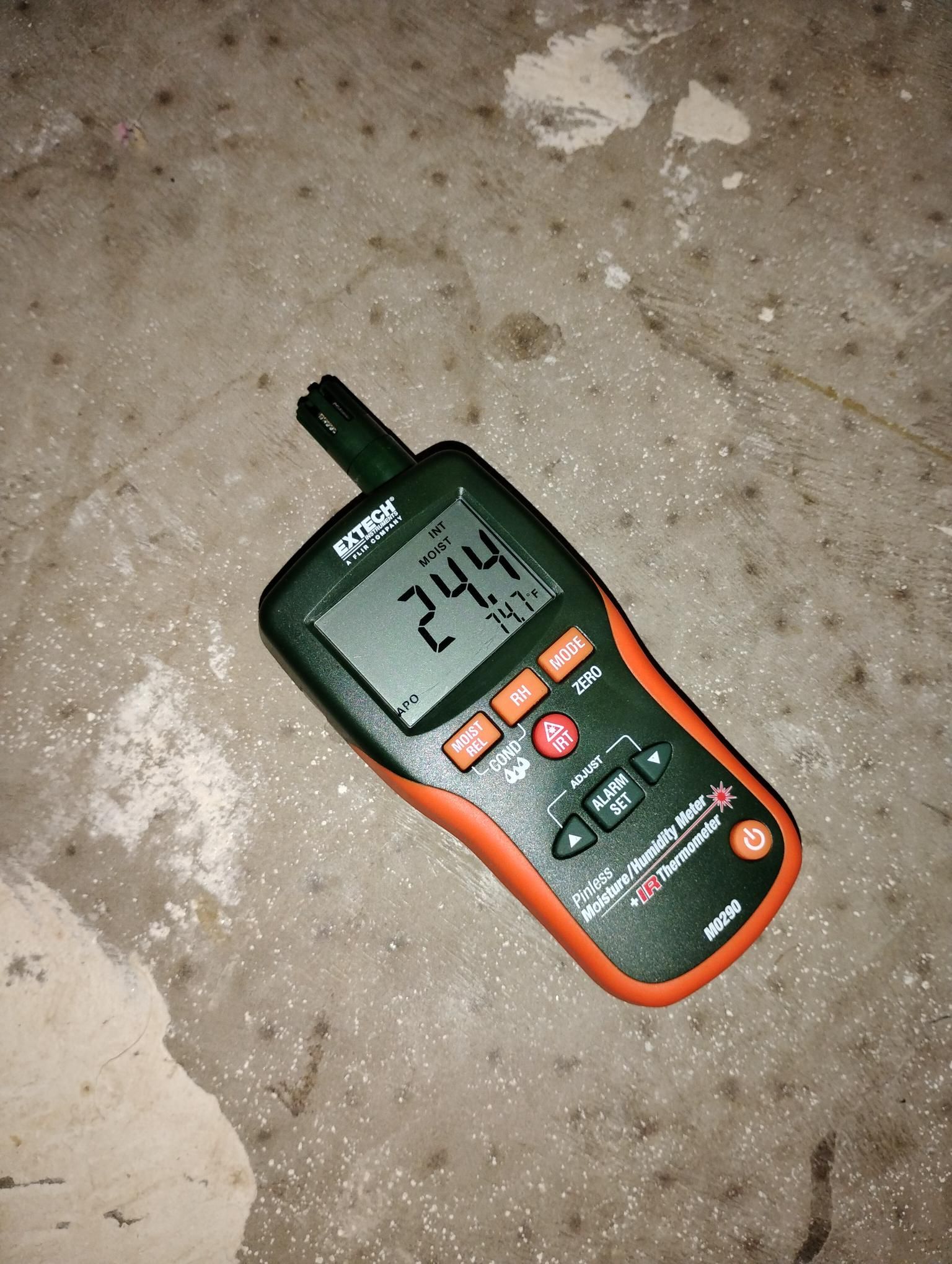 A moisture meter on a concrete surface displays 24.4 and 74.7. The device has an orange and green design.