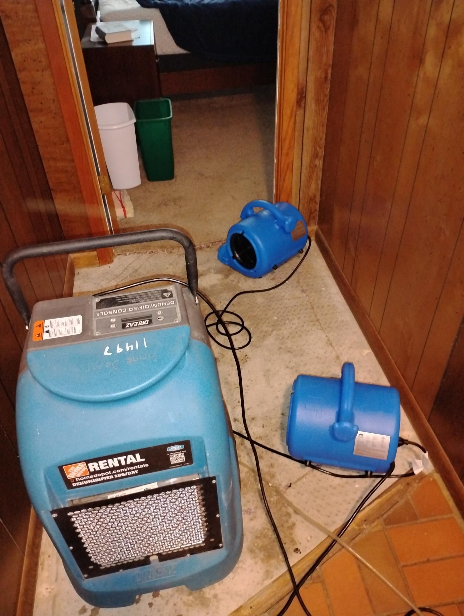 Two blue dehumidifiers and fans are drying a flooded room with wood paneling.