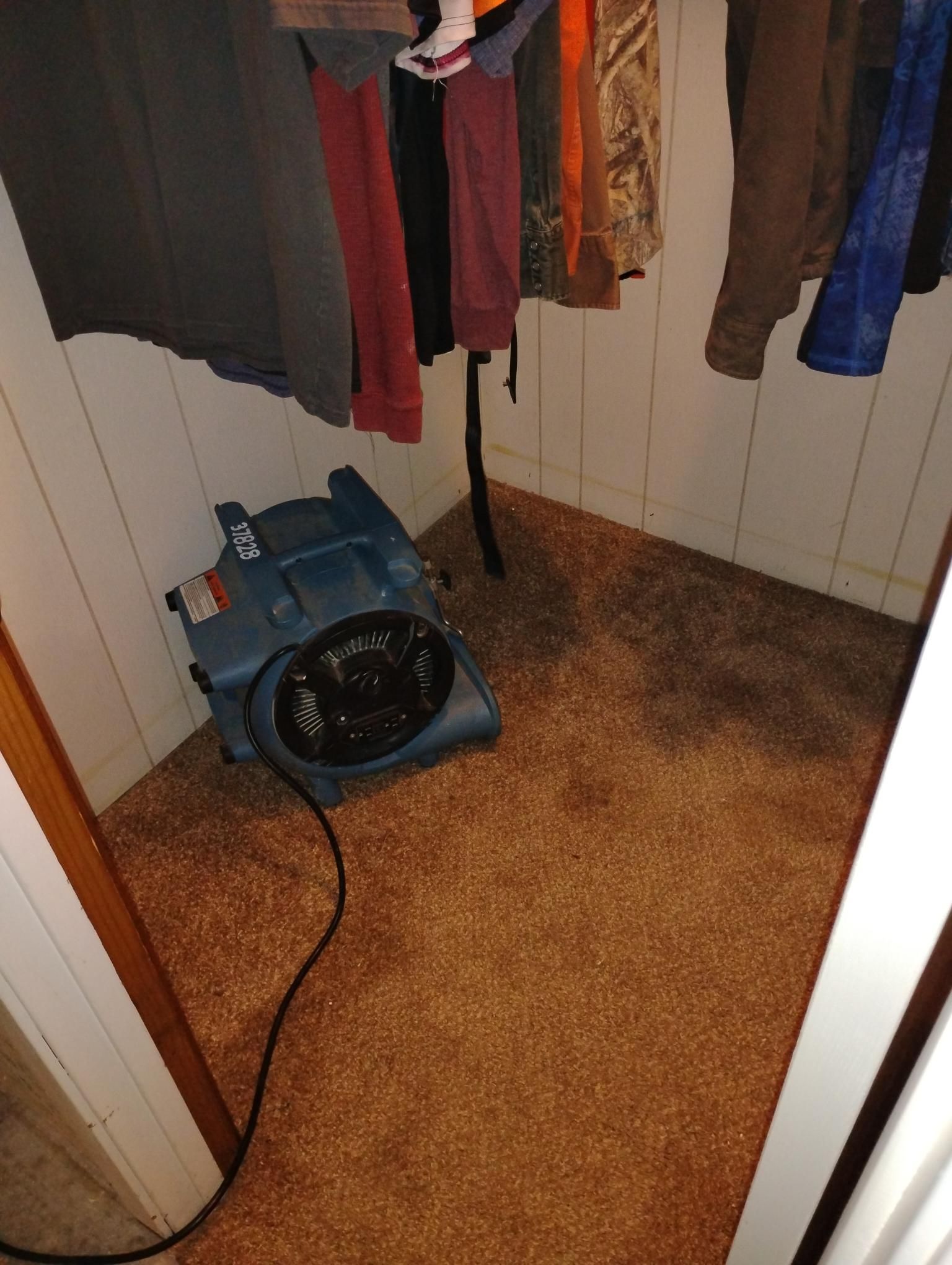 Blue dehumidifier in a closet with wet carpet and hanging clothes.