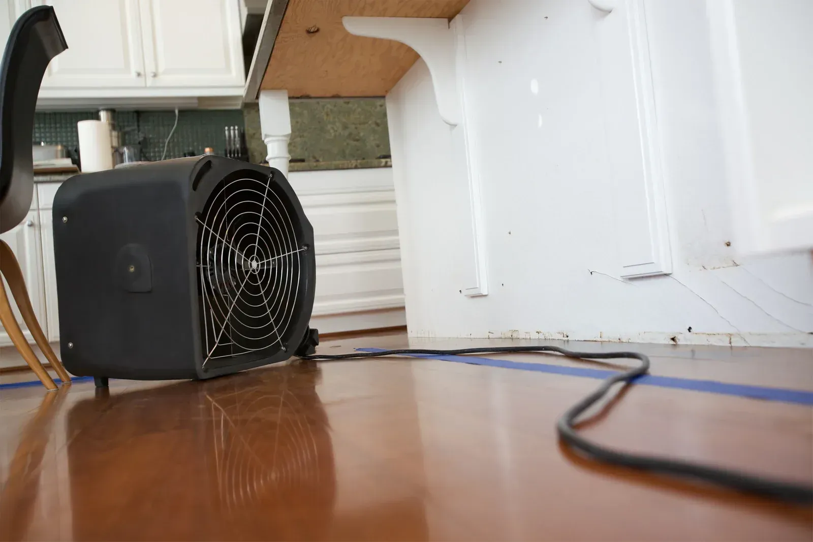 Black fan on a brown floor, next to a white wall with blue tape; in a kitchen.