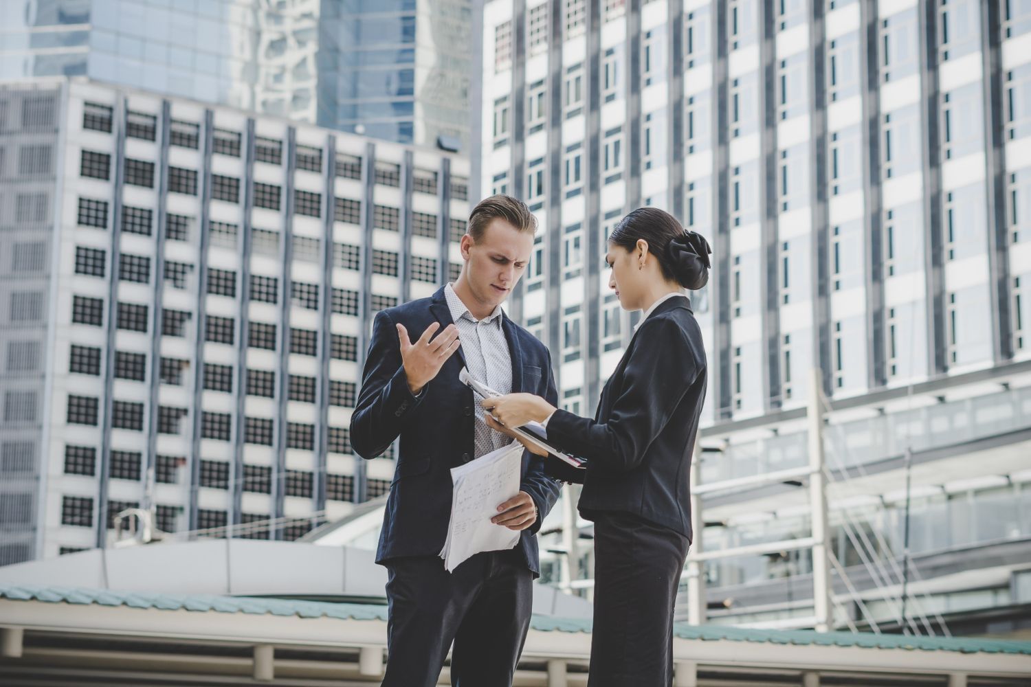 Two business professionals in suits reviewing documents outdoors in front of skyscrapers; man gesturing, woman looking at papers.
