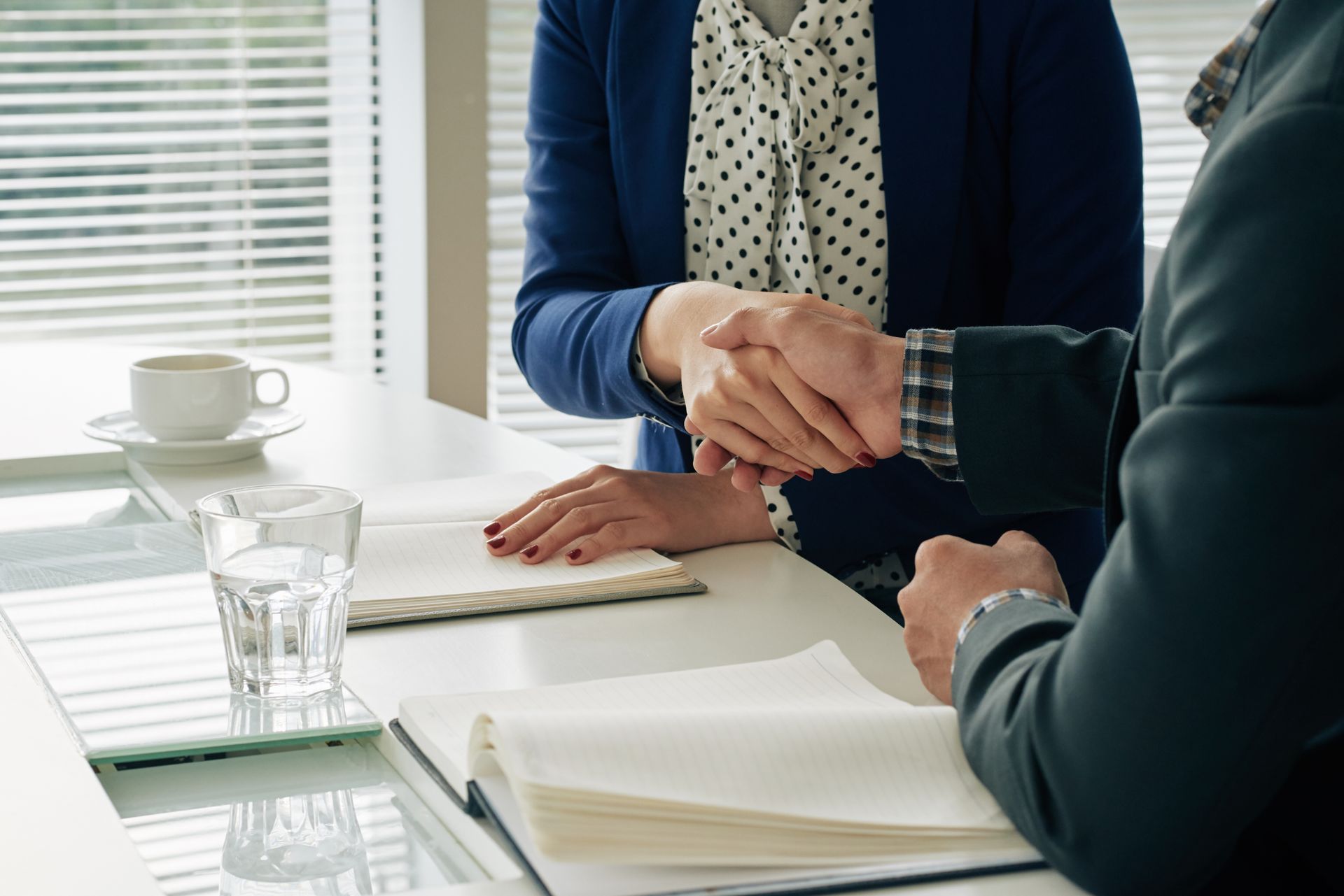 Two people in business attire shake hands over a table with open notebooks, a water glass, and a coffee cup, in an office setting.