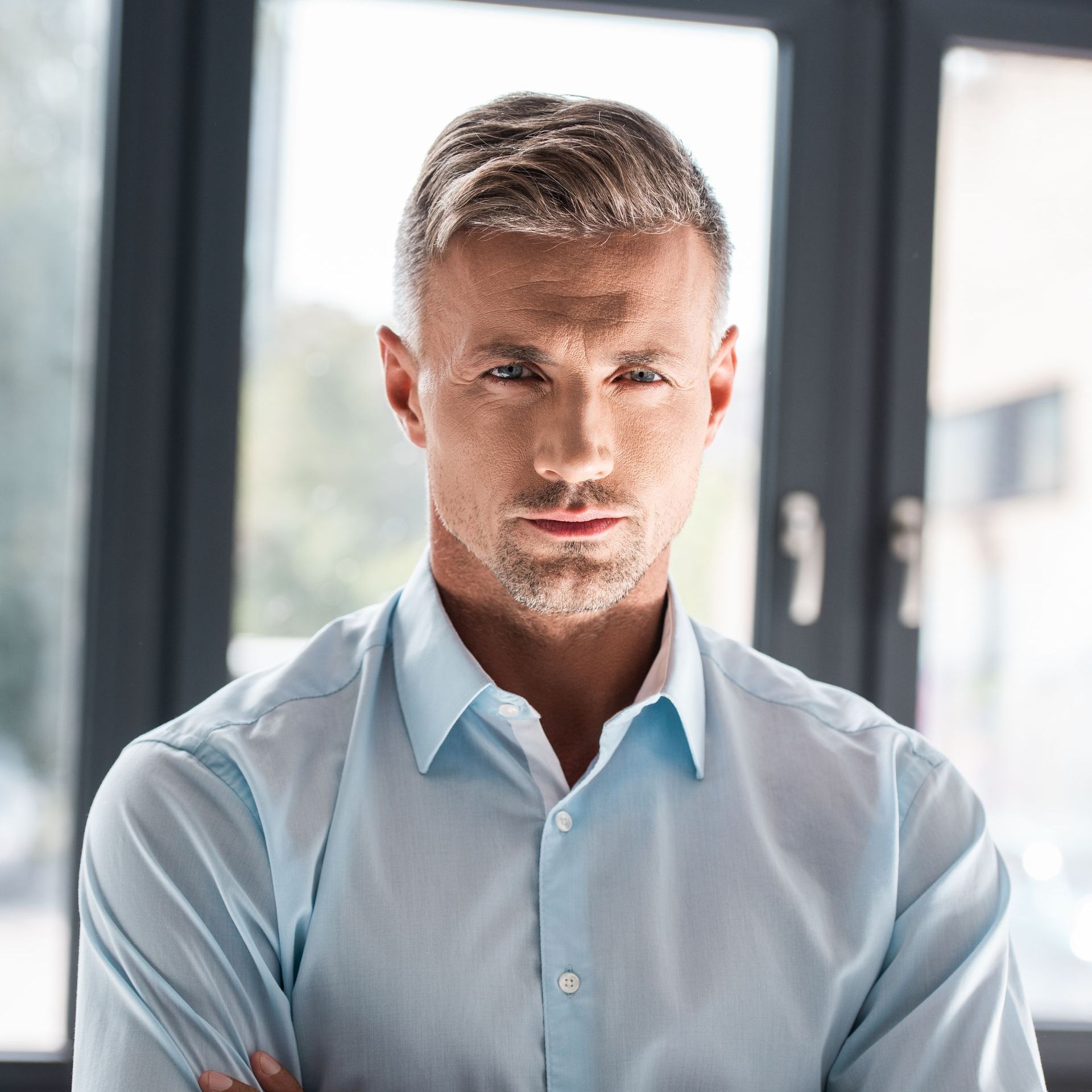 A man in a light blue shirt is standing in front of a window with his arms crossed.
