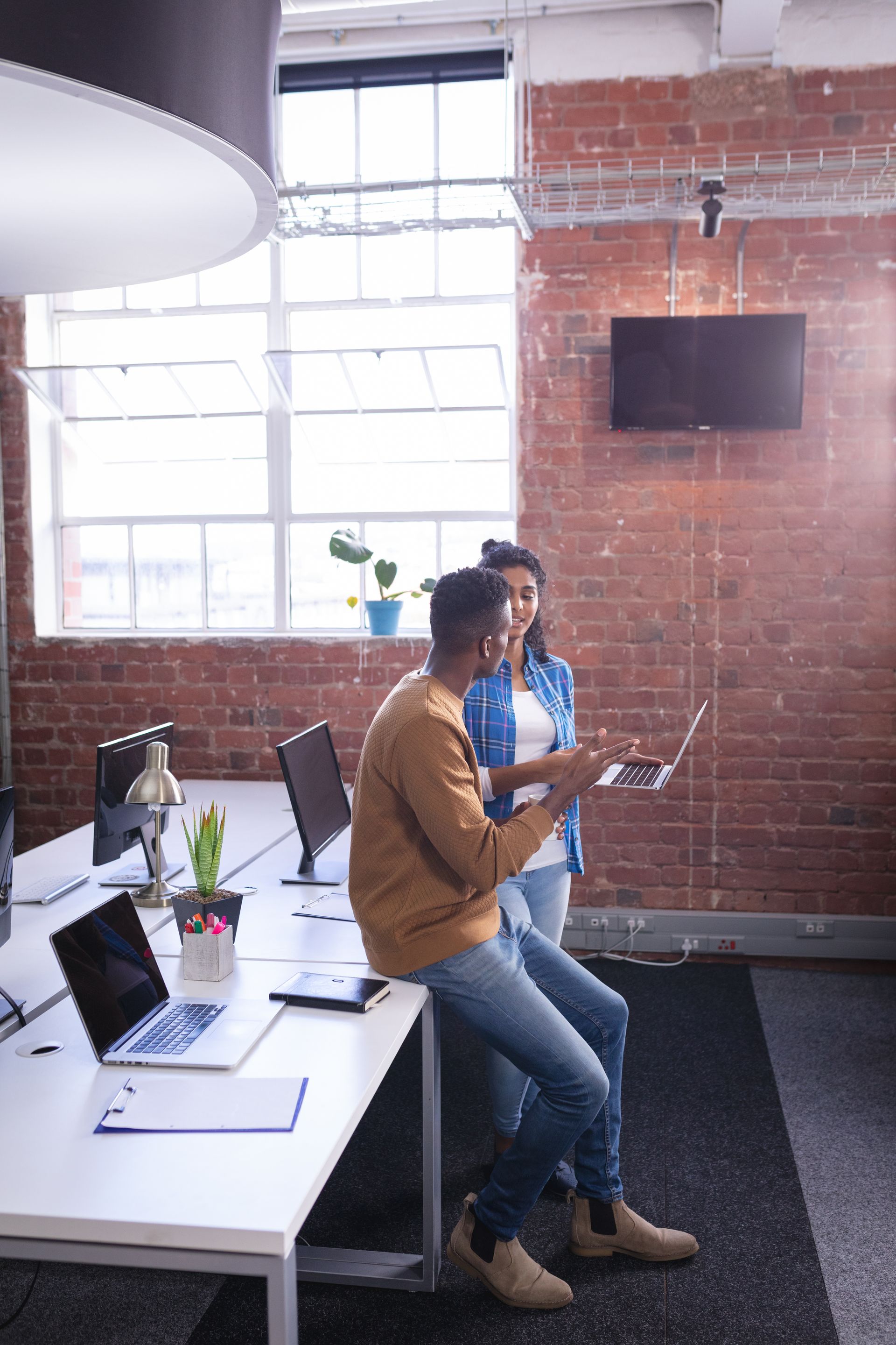 Two colleagues at a desk in an office setting, looking at a laptop. One is seated, the other standing, both gesturing.