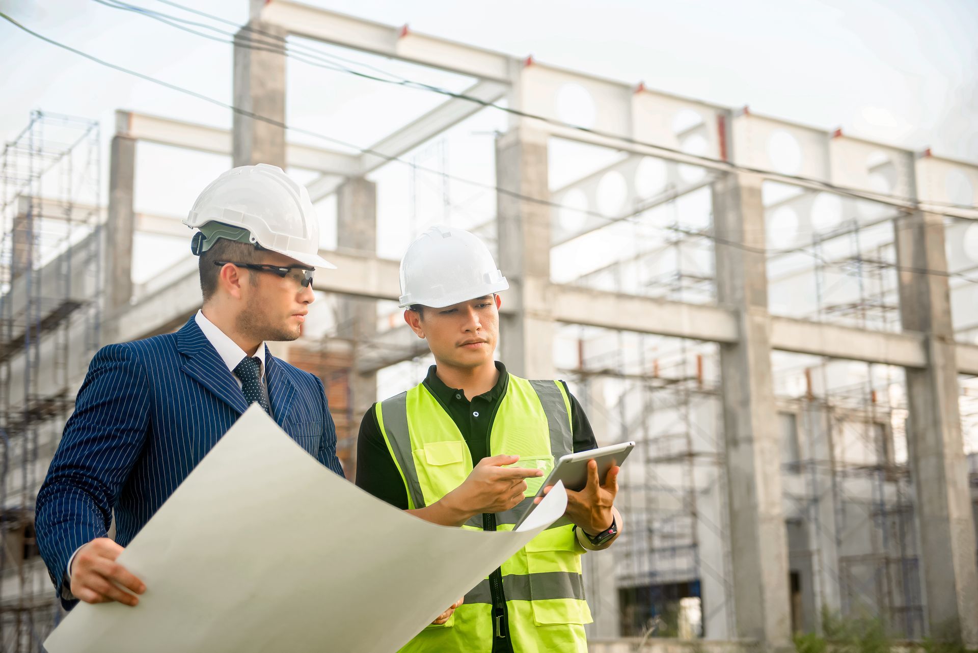 Two construction workers, one in a suit, the other in a vest, reviewing blueprints at a construction site with the steel frame of a building in the background.