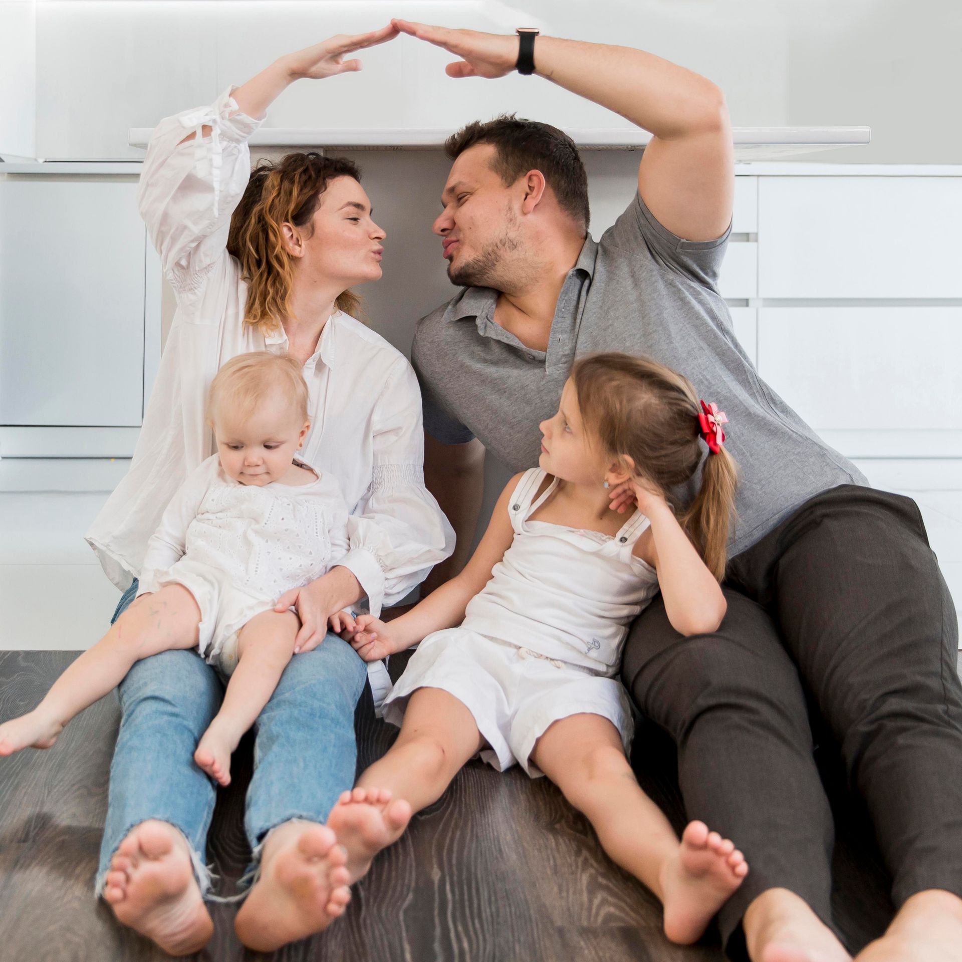 Family of four sitting on the floor making a roof with their hands, smiling and looking at each other.