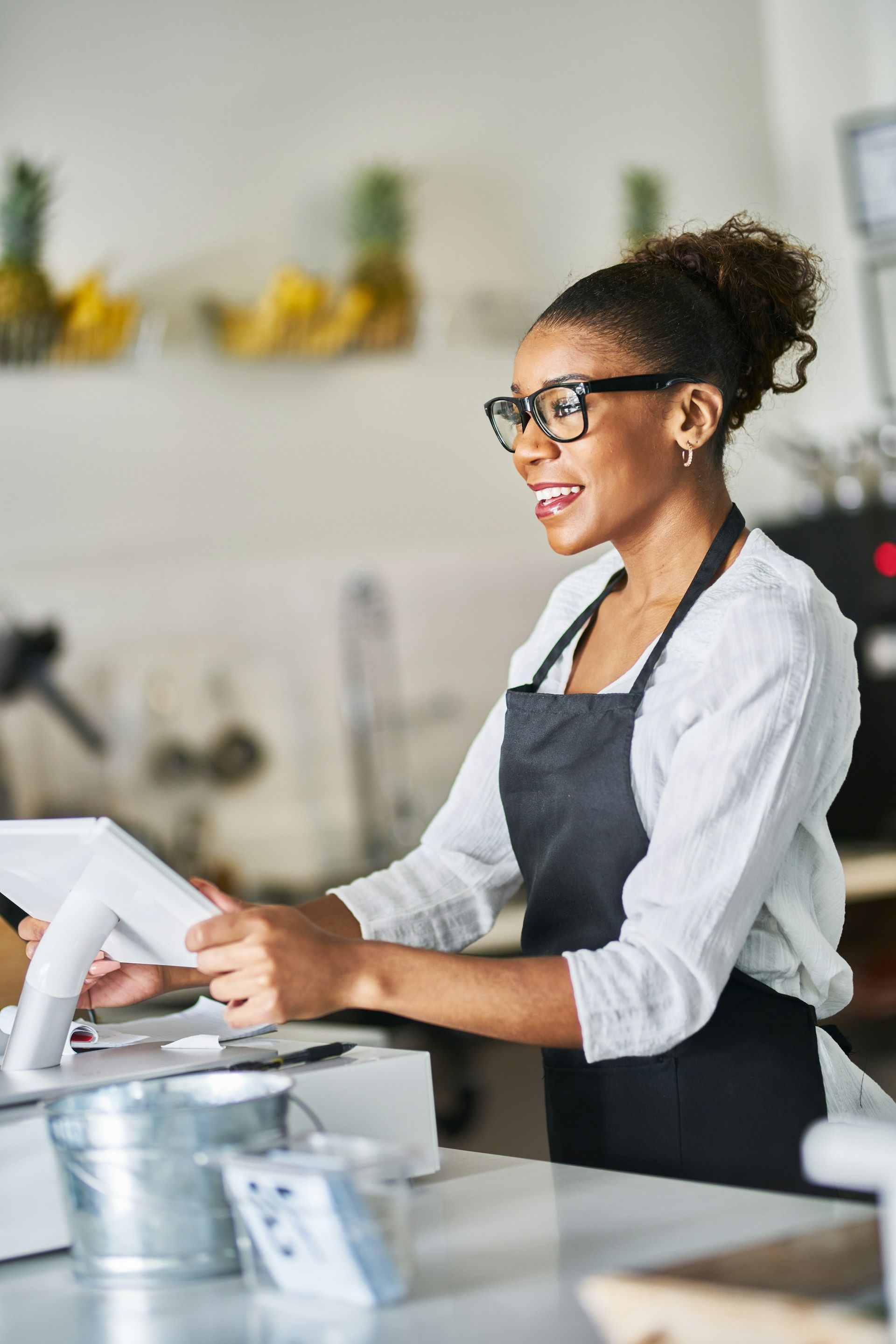 Woman with glasses and a dark apron smiles while operating a point-of-sale system at a cafe counter.