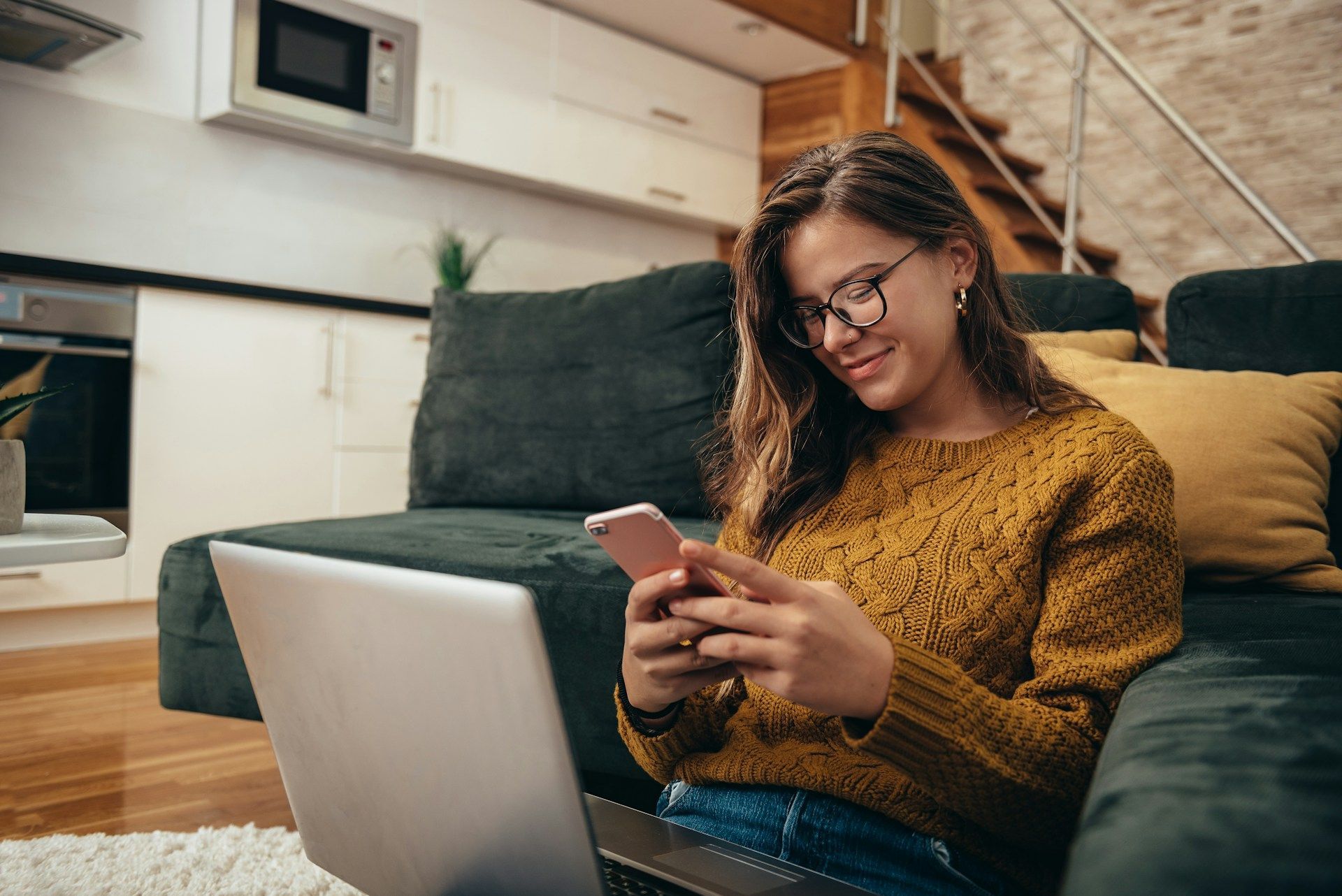 Woman with glasses, smiling, using a phone, sitting with a laptop in a living room with a dark green couch.