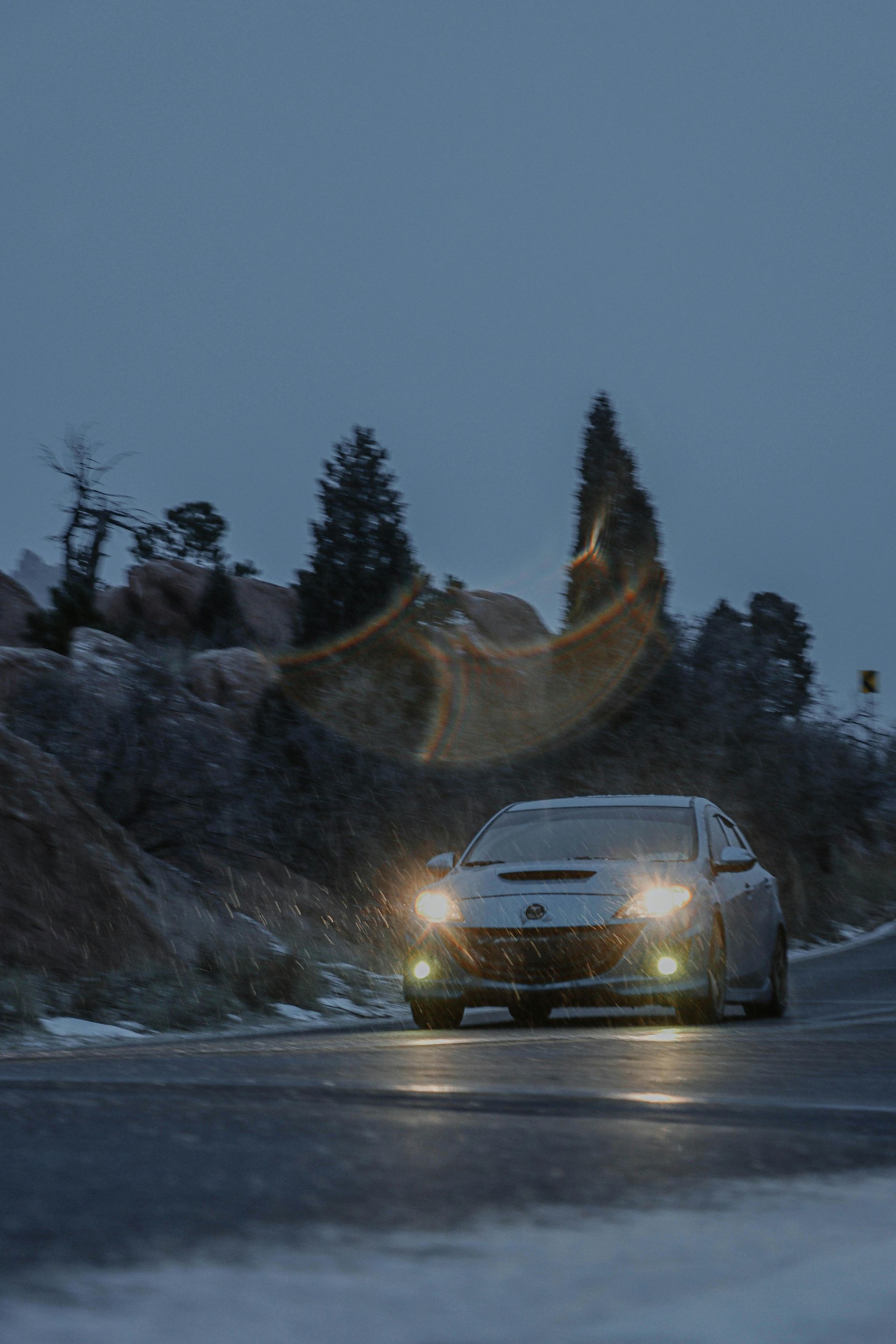 A silver car drives on a snowy road at dusk, headlights on, mountain backdrop.