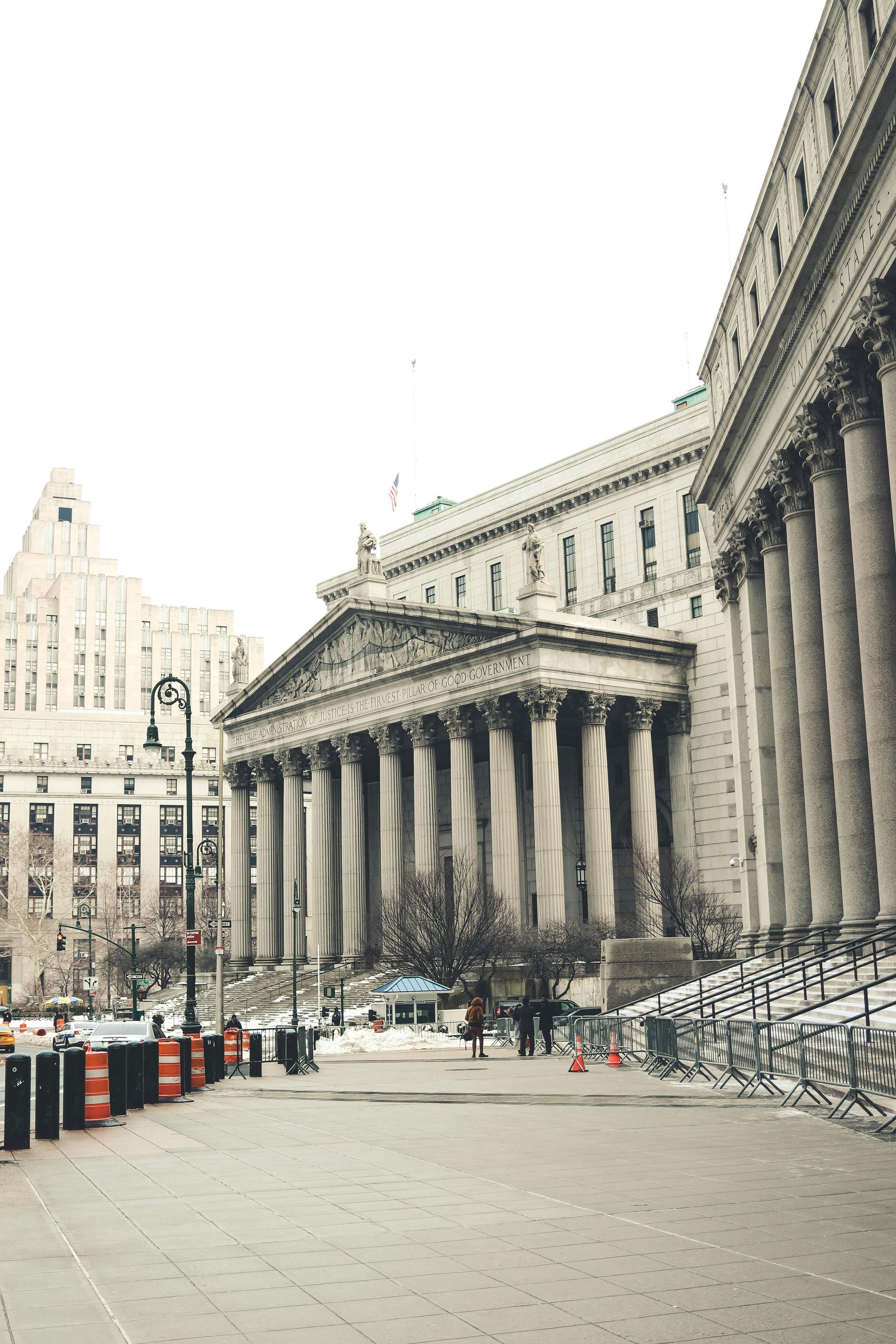 Classical architecture building with large columns and a long facade. Sidewalk in front with other buildings visible in the background, overcast sky.