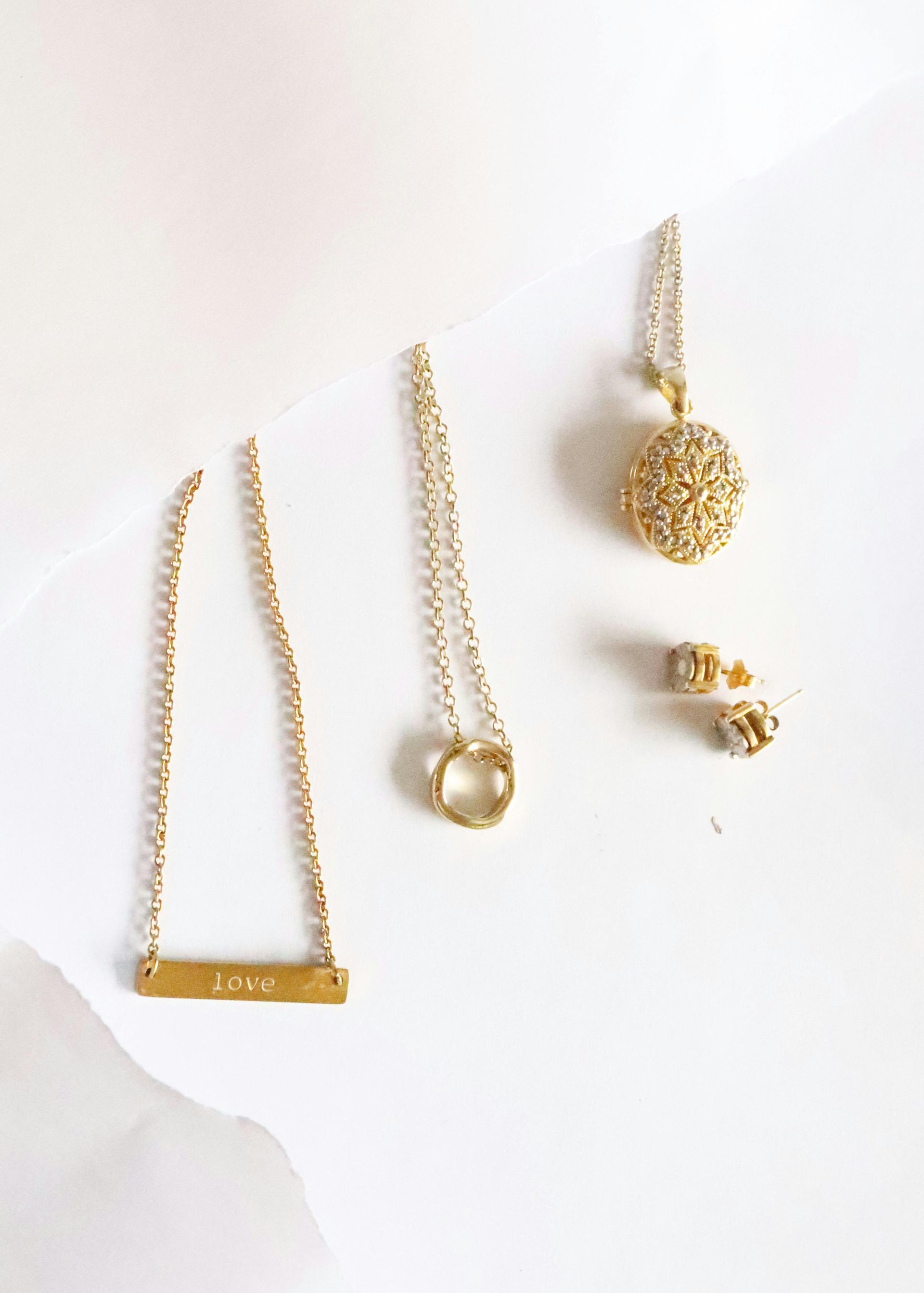 Gold jewelry set on a white background. Includes necklaces with a bar, ring, and sphere pendant, plus small stud earrings.