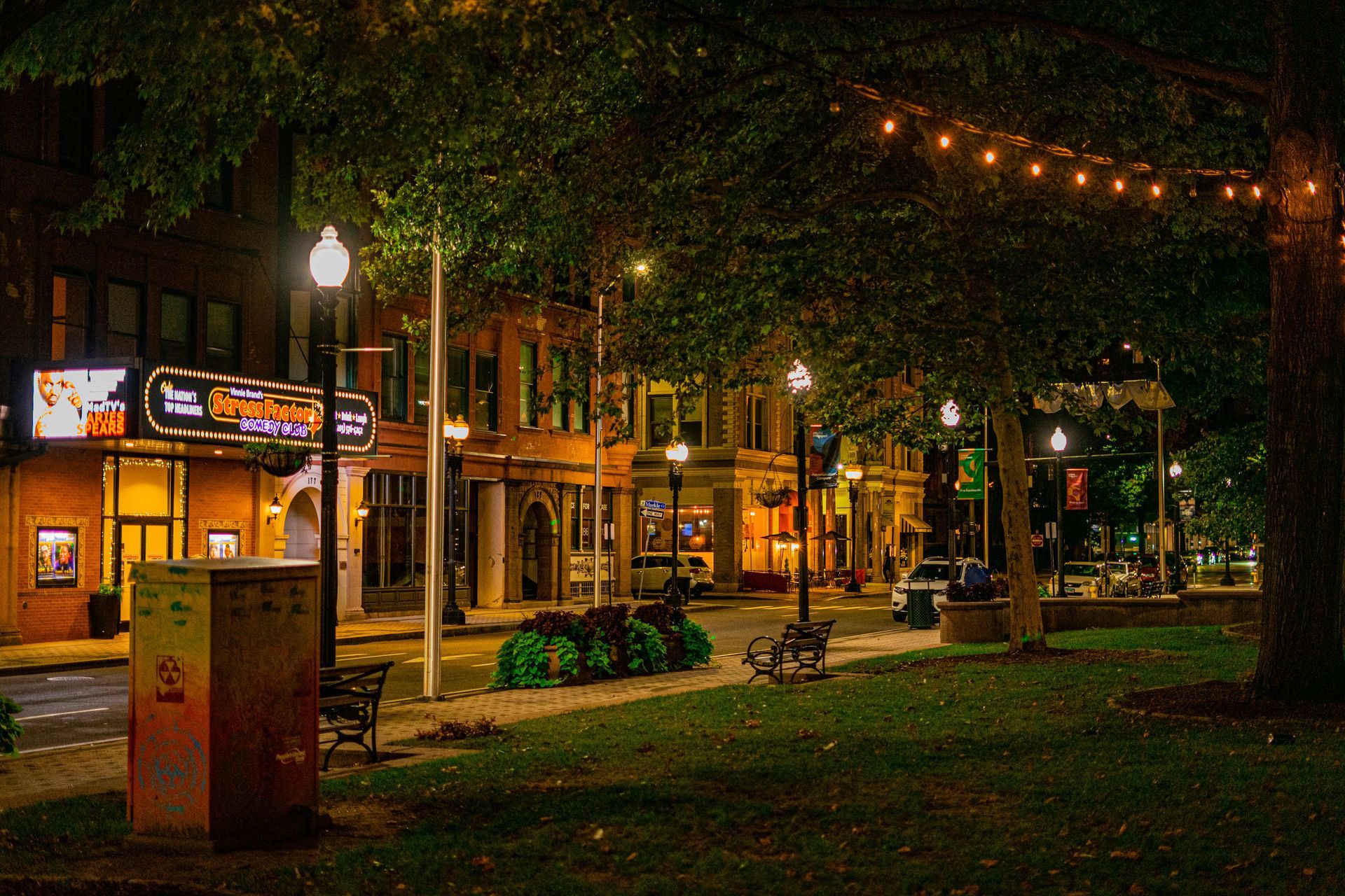 Nighttime street scene with trees, benches, and buildings lit by streetlights and string lights. A theater with illuminated signage is on the left.