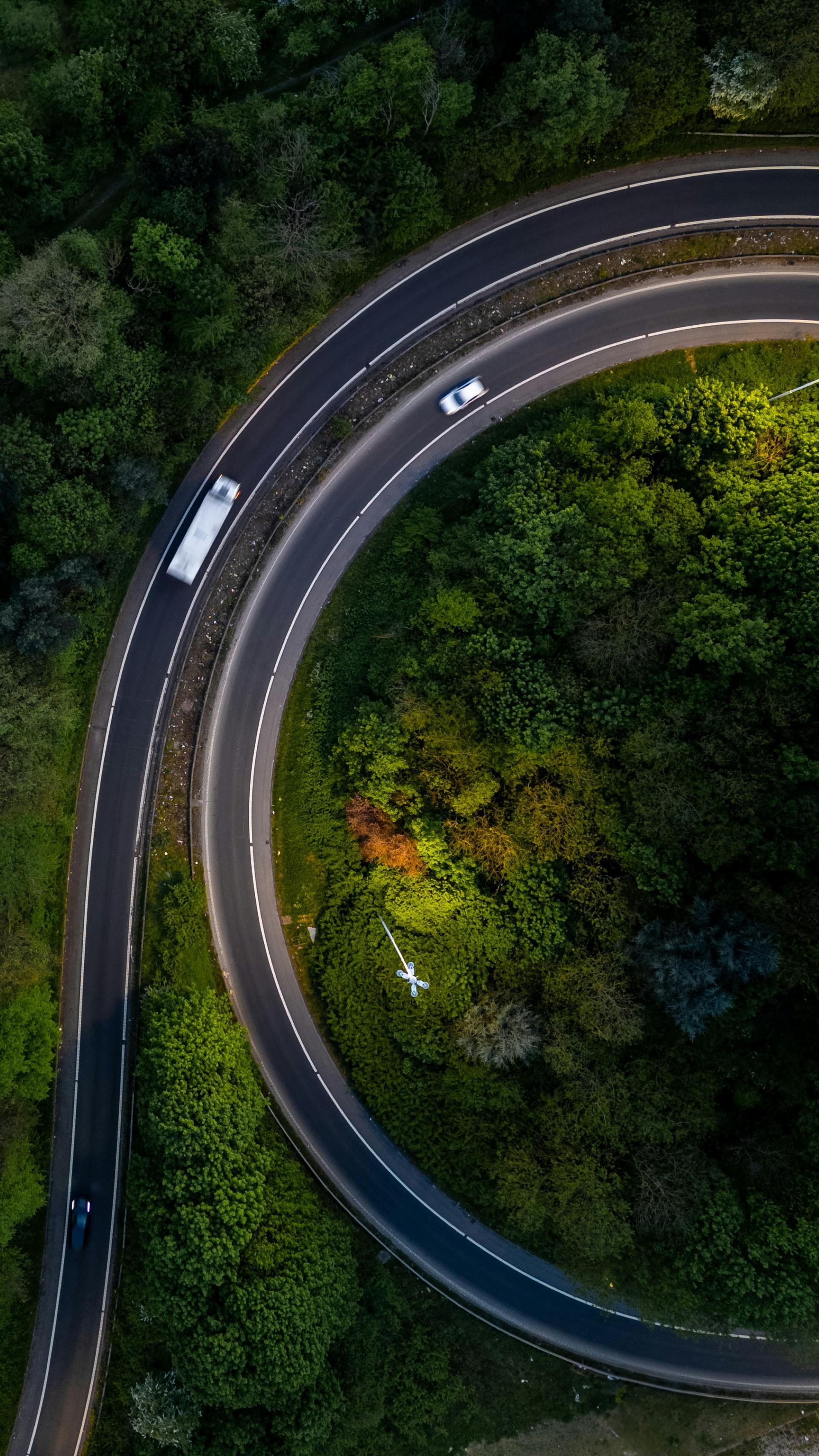 Curved road winding through lush green forest; a white truck and car are visible.