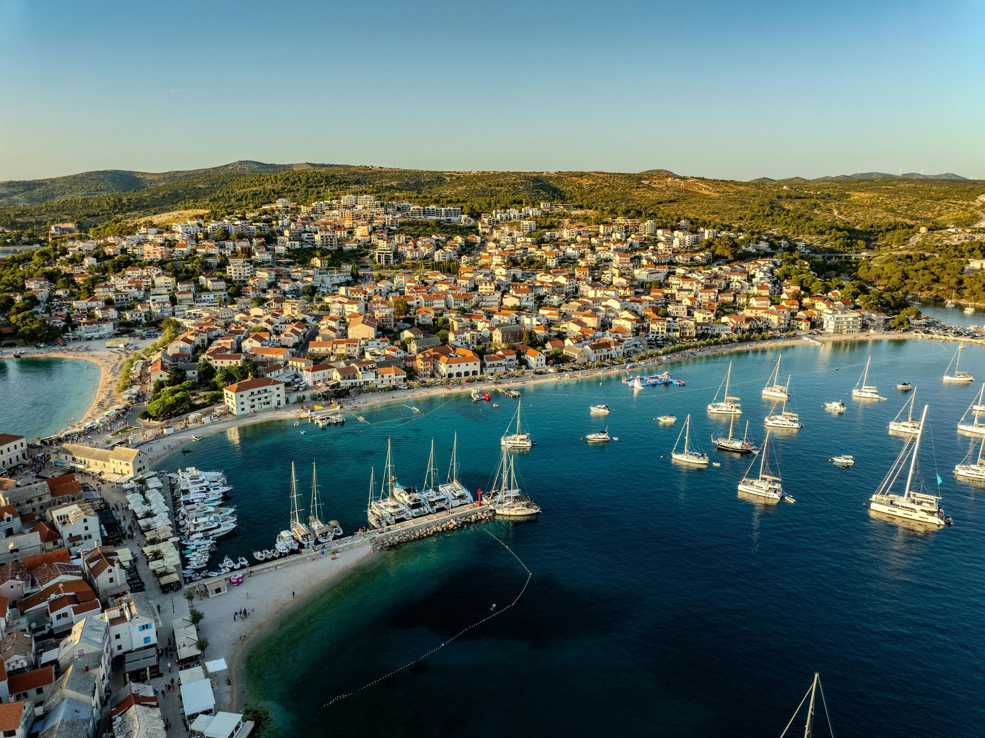 Aerial view of a coastal town with buildings along a beach, boats in the turquoise water, and hills in the background.
