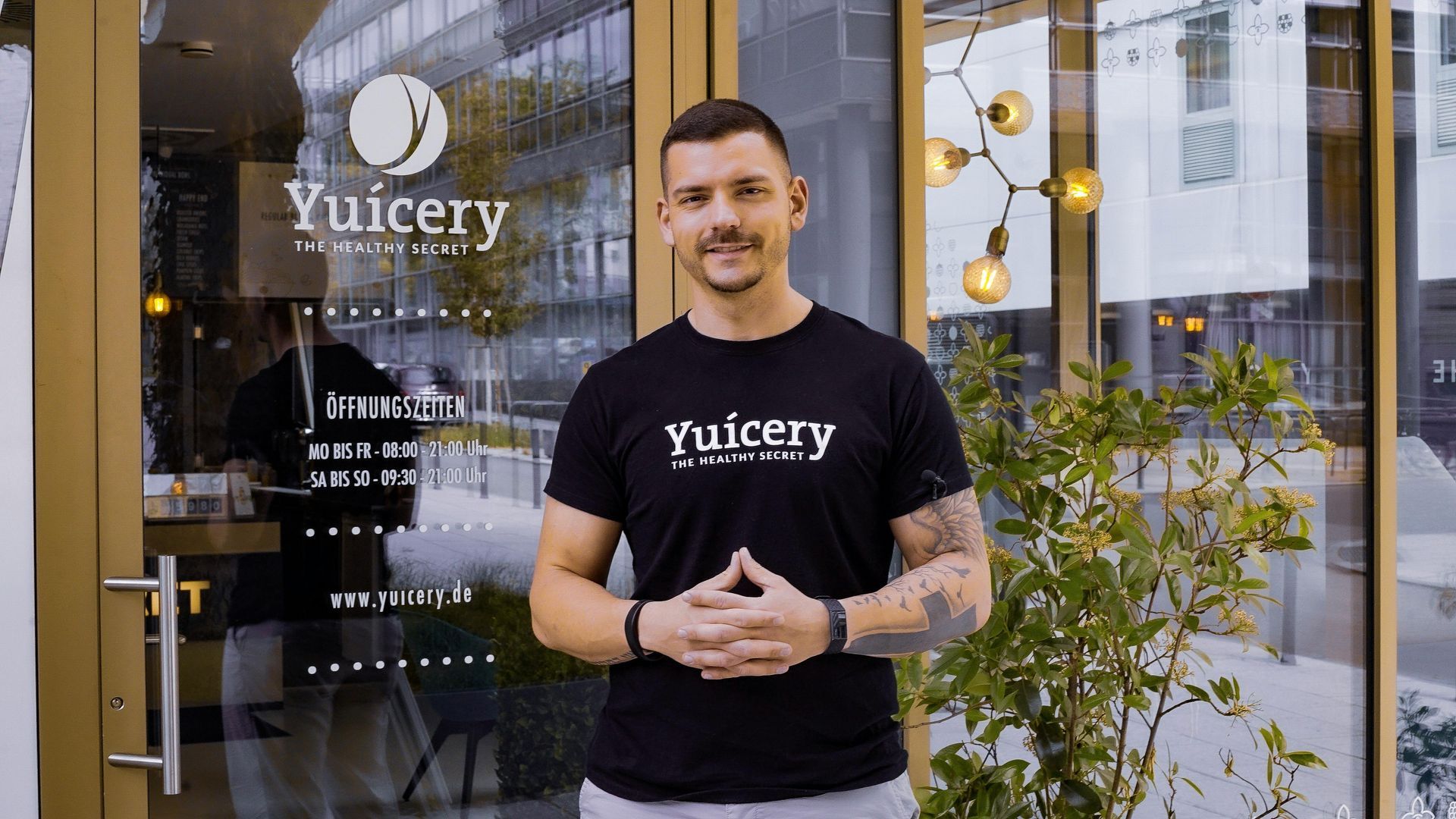 A man with a mustache stands in front of a juice bar called Yuicery. He wears a black t-shirt, and his hands are clasped.
