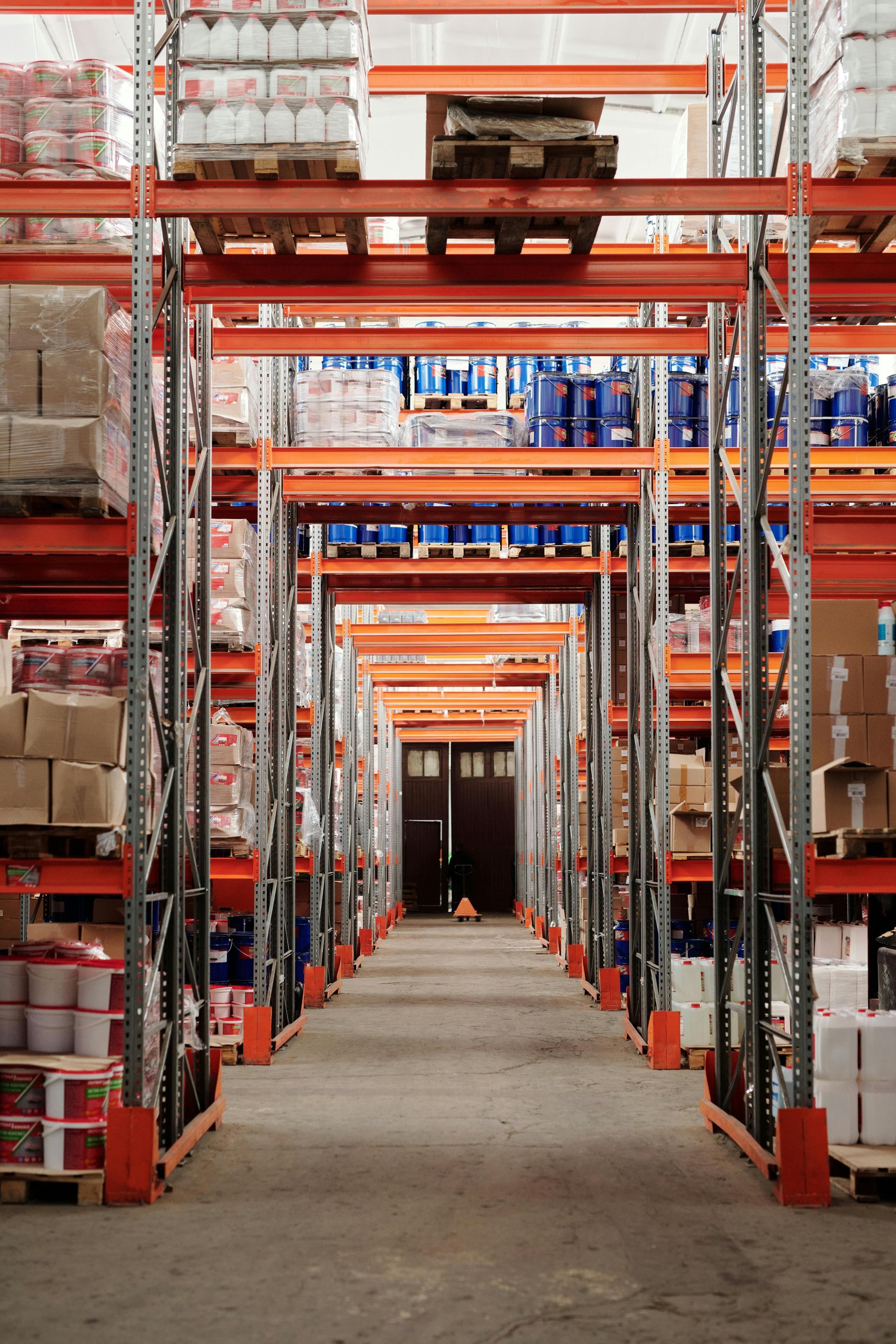 Rows of tall, orange shelving filled with packaged goods in a warehouse; a long, empty aisle leads to a door.