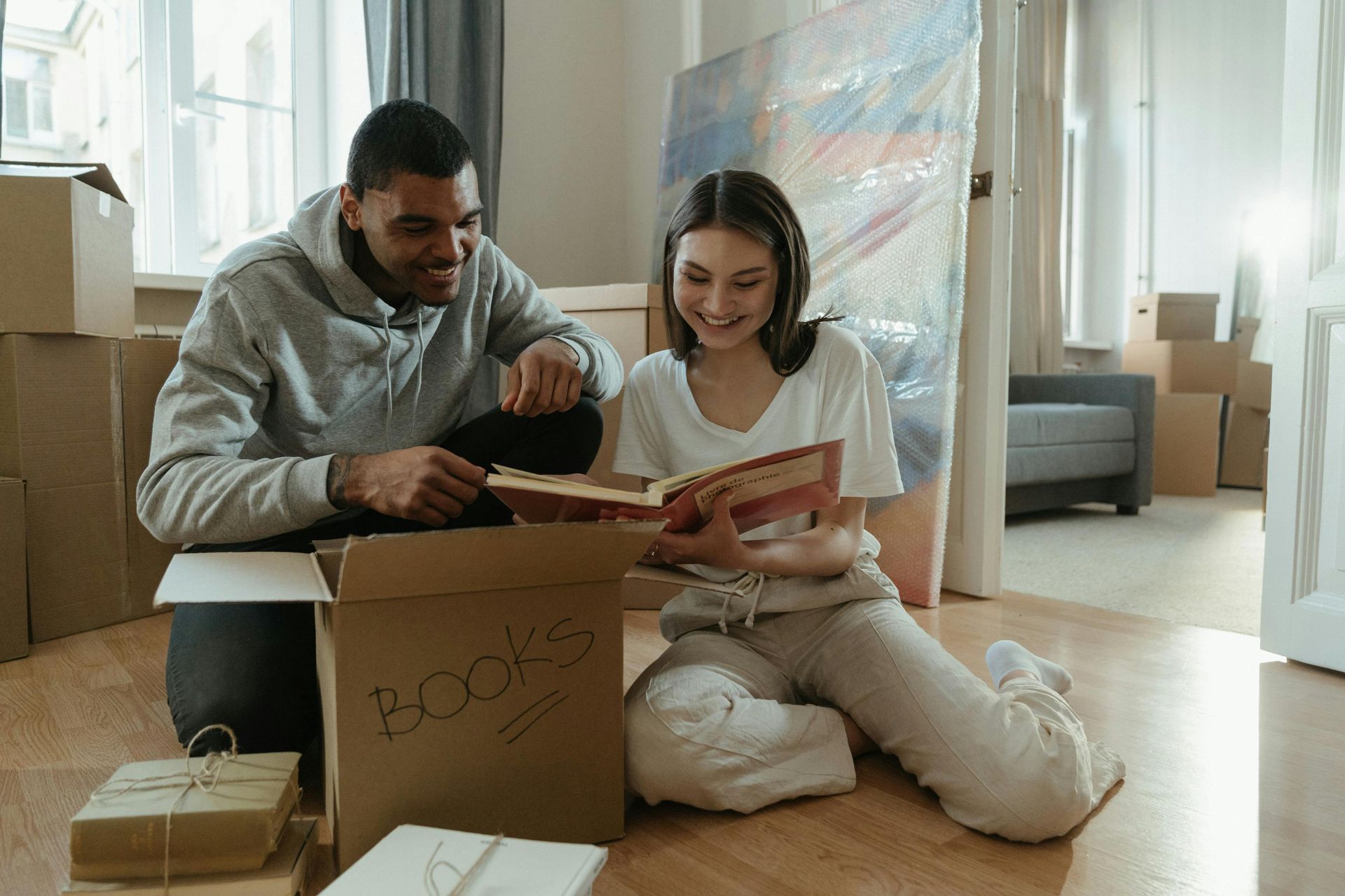 Couple unpacking boxes in a new home, looking at a book together. Man smiles, woman reads with joy.