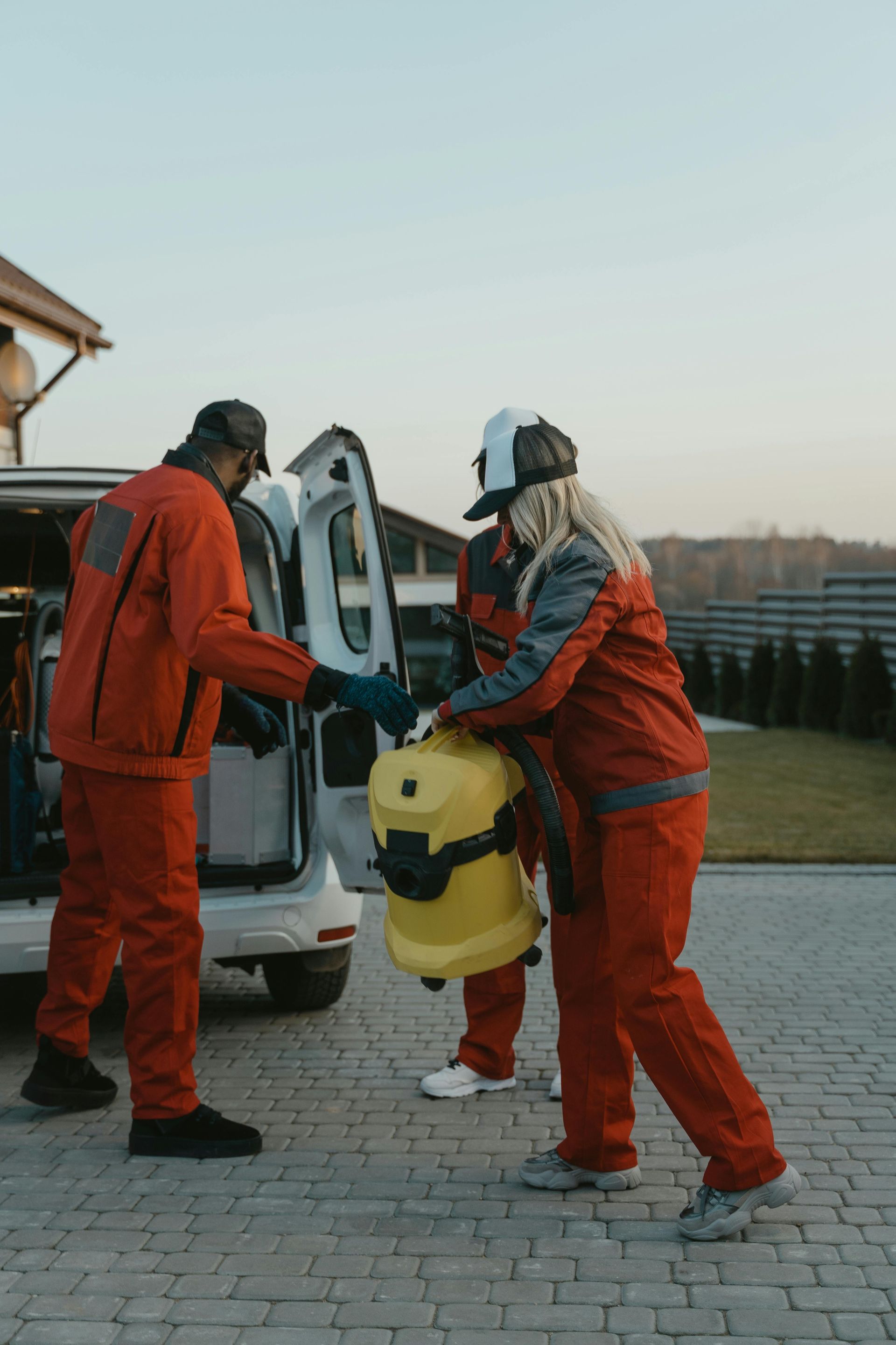 Three people in red coveralls load a yellow vacuum cleaner into the back of a white van parked on a paved driveway.