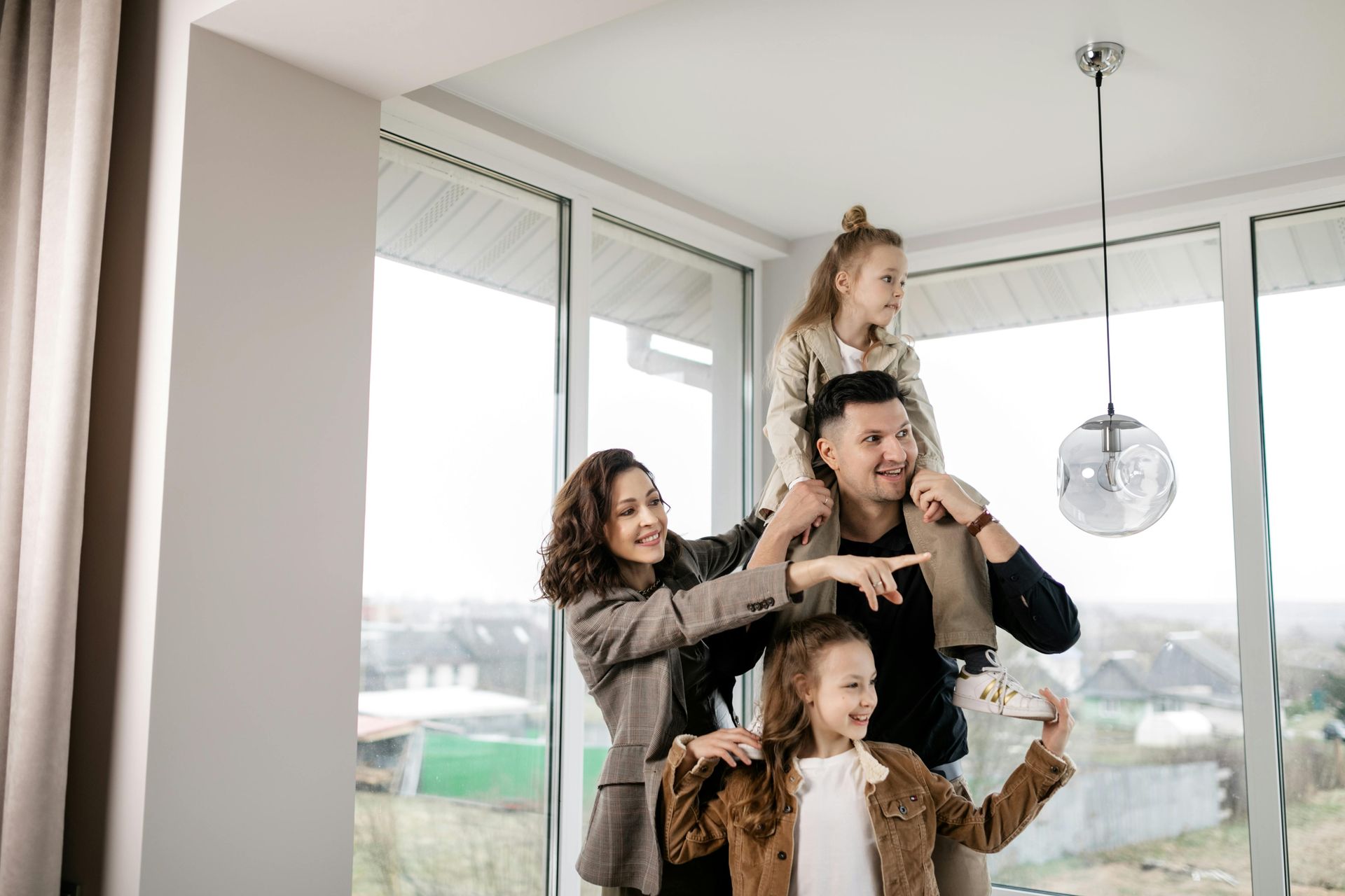 Family of four in modern home, with a girl on the father's shoulders and another child in front.  Smiling, looking at the camera.