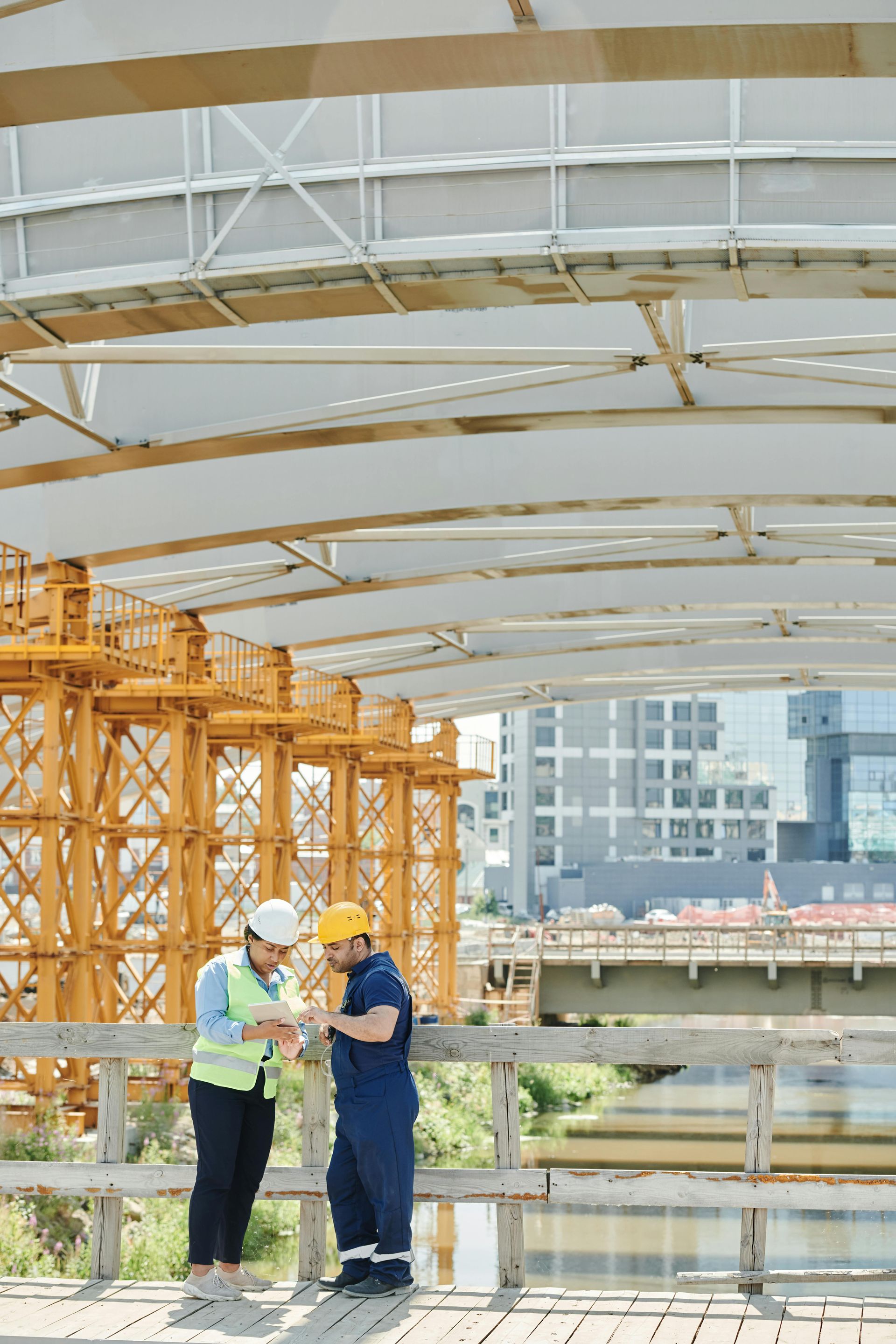 Two construction workers in helmets review documents on a wooden platform under a bridge, with machinery in the background.