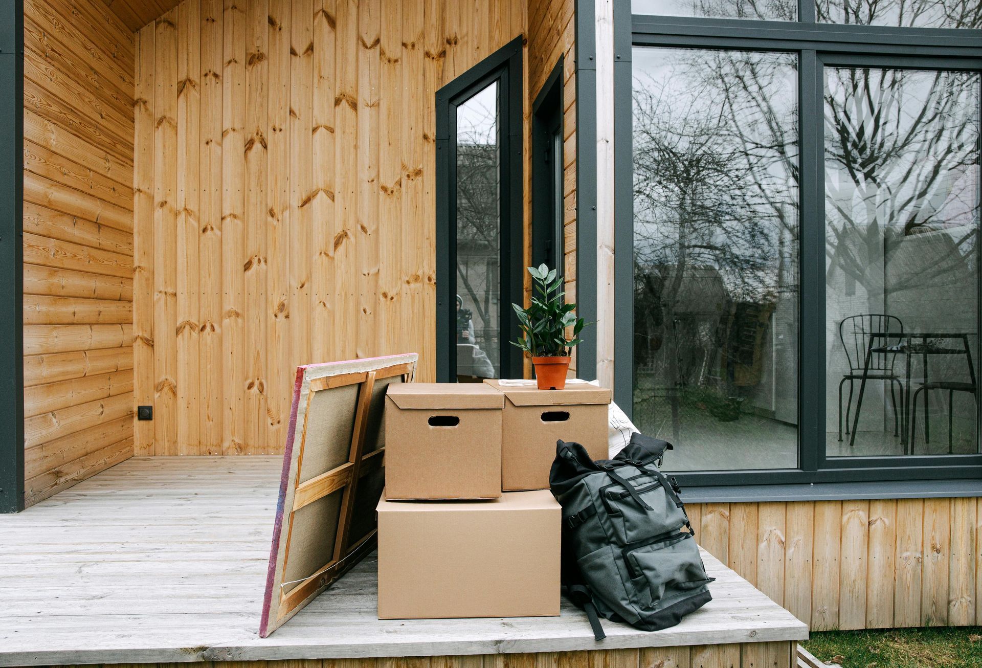 Moving boxes, backpack, and artwork on a wooden porch outside a wood-clad building with large windows.