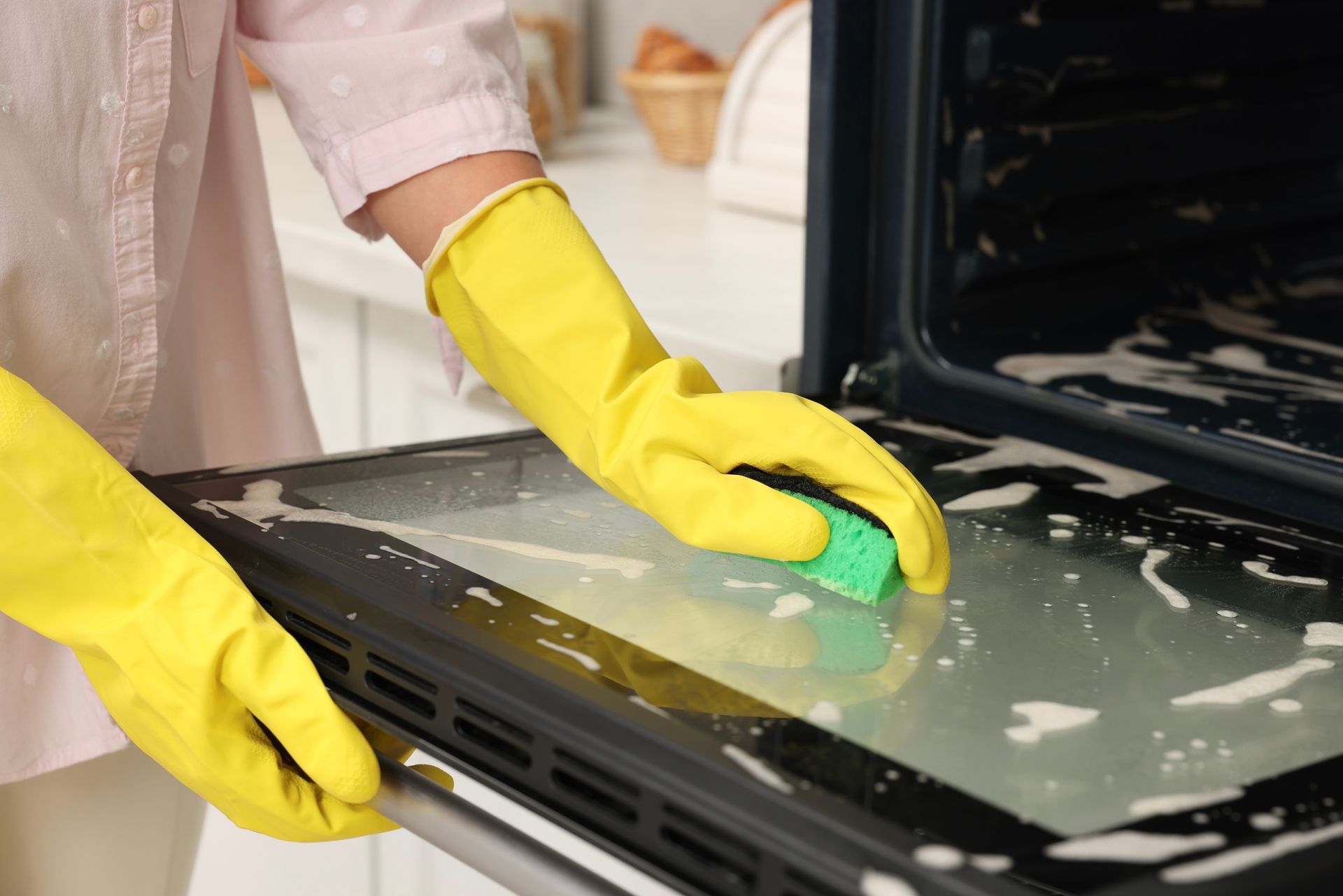 Person wearing yellow gloves cleans an oven door with a sponge.