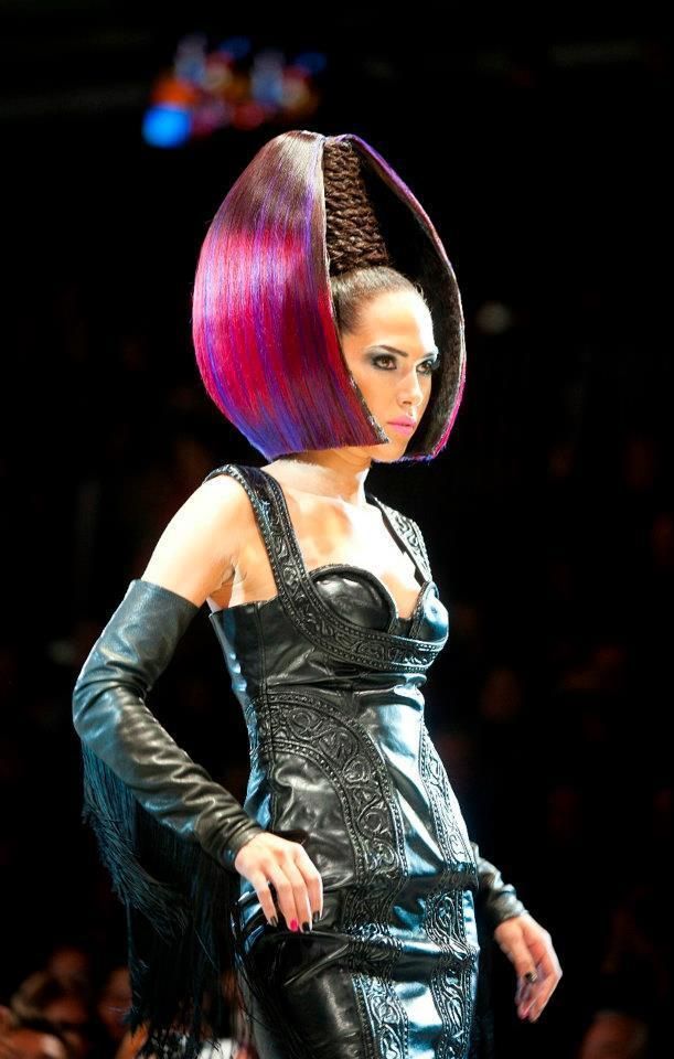Model in a black latex dress and long gloves posing on a runway with a tall purple-and-pink sculptural hairstyle