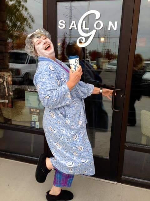 Person in a blue patterned robe laughing outside a salon, holding a drink and leaning against the door