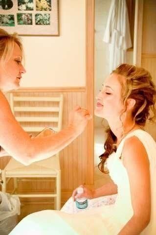 Two women in a bathroom, one applying makeup to the other as she sits in a white dress.