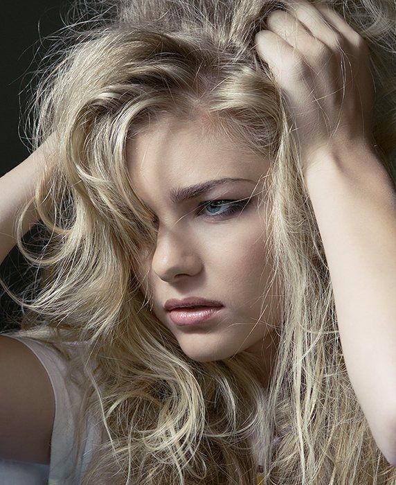 Close-up portrait of a blonde woman with tousled hair and a pensive expression, resting her hand on her head