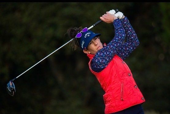 Woman in a red vest and patterned shirt swings a golf club outdoors. She wears sunglasses and a visor.