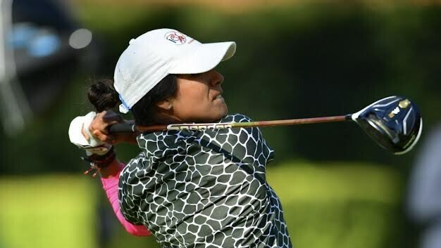 Golfer swings a club on a course, wearing a white cap, patterned shirt, and pink arm sleeves.
