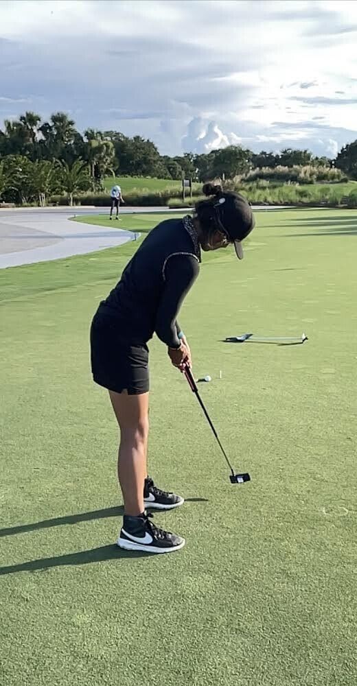 A person in black attire putts a golf ball on a green course, with a hat on and mountains in the background.