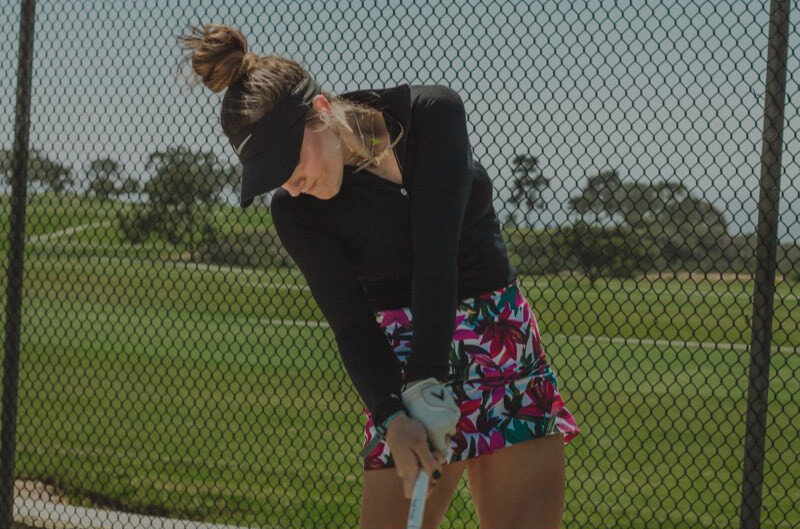 Woman in golf attire swinging a club on a driving range, wearing a visor and floral skirt.