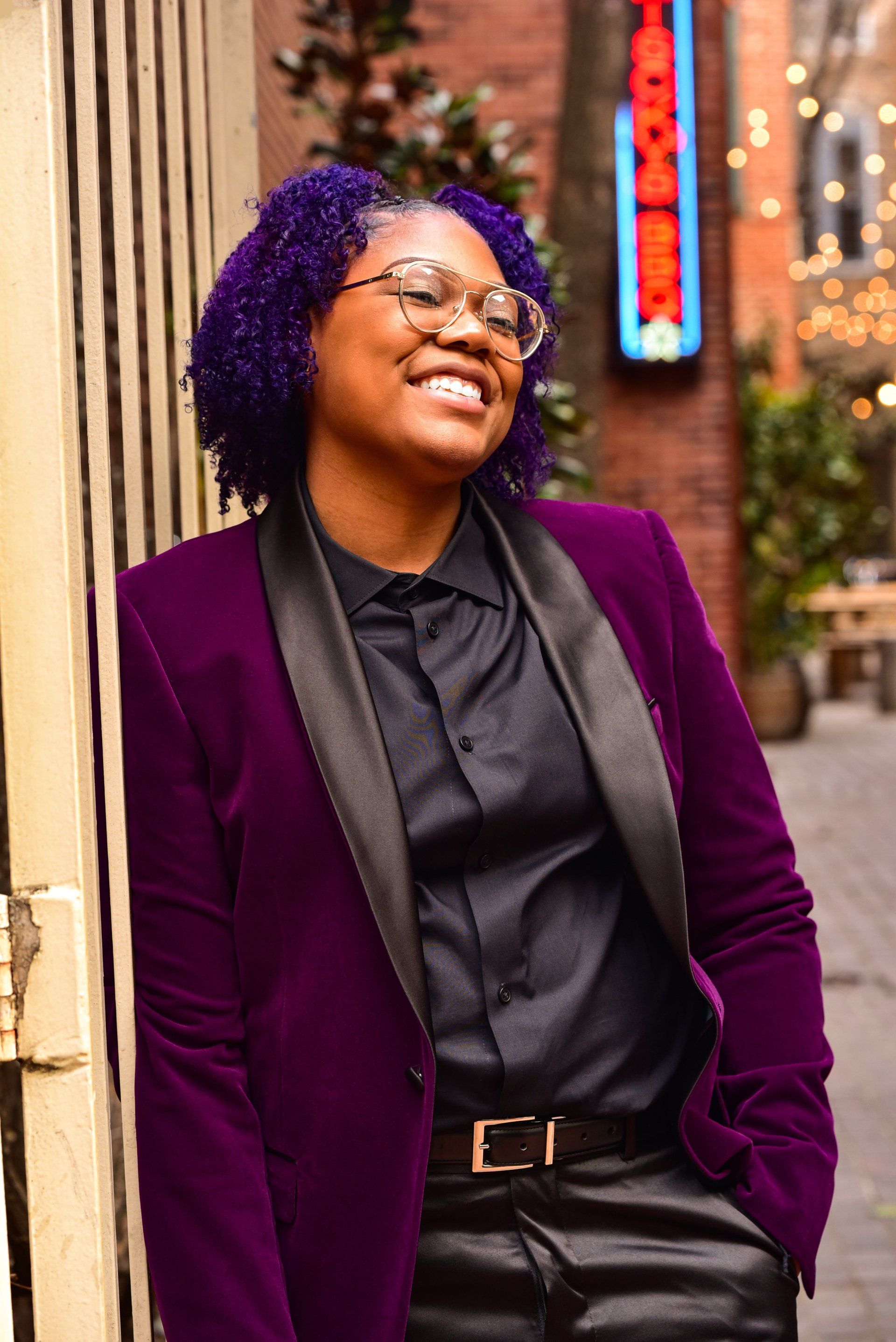 a woman with purple hair and glasses is wearing a purple jacket and black shirt .