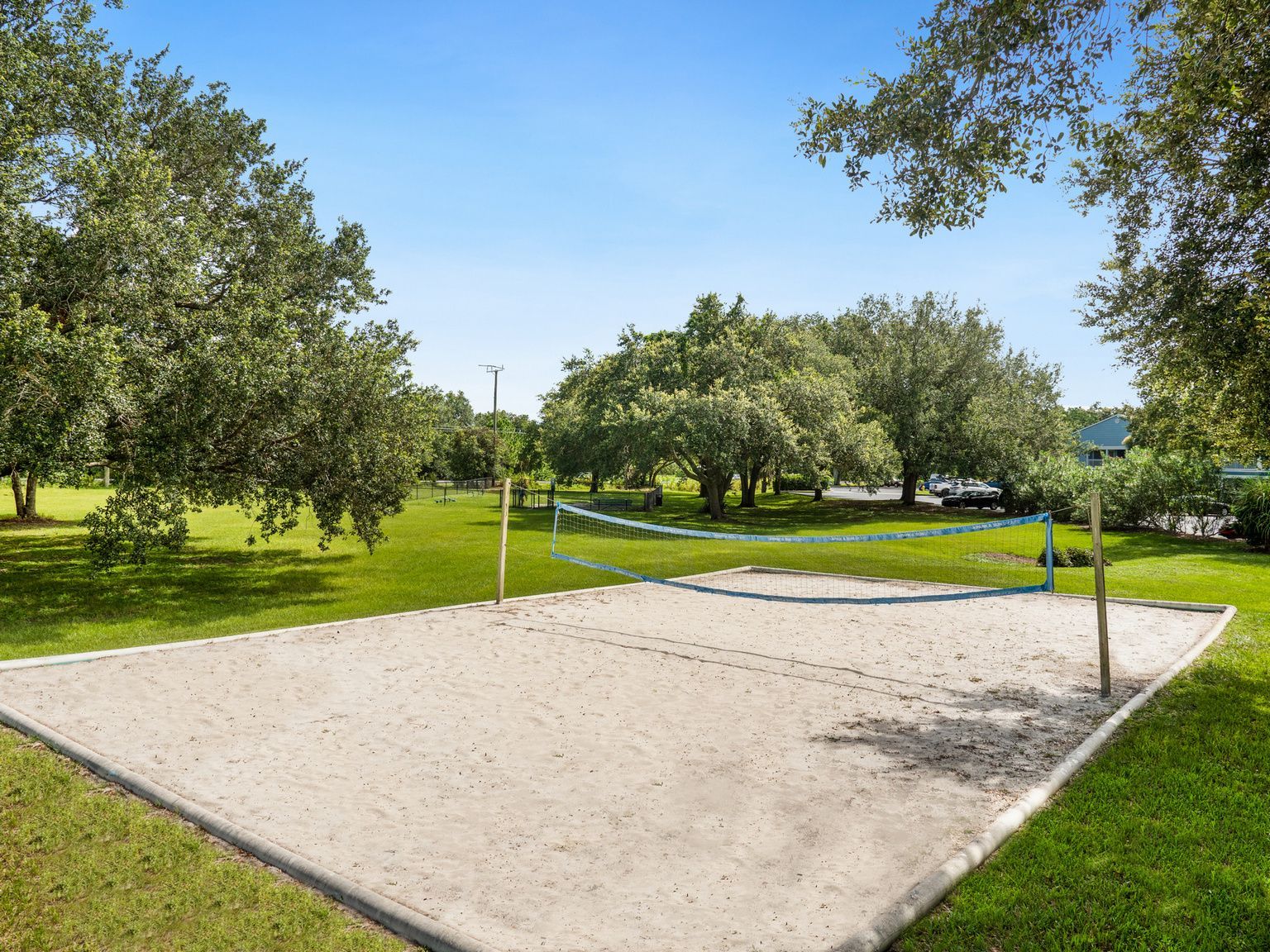 Sand volleyball court with net, surrounded by green grass and trees under a blue sky.