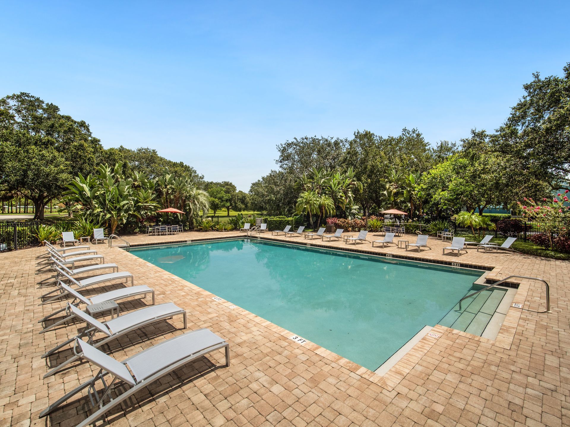 Pool with lounge chairs, brick patio, surrounded by trees under a blue sky.