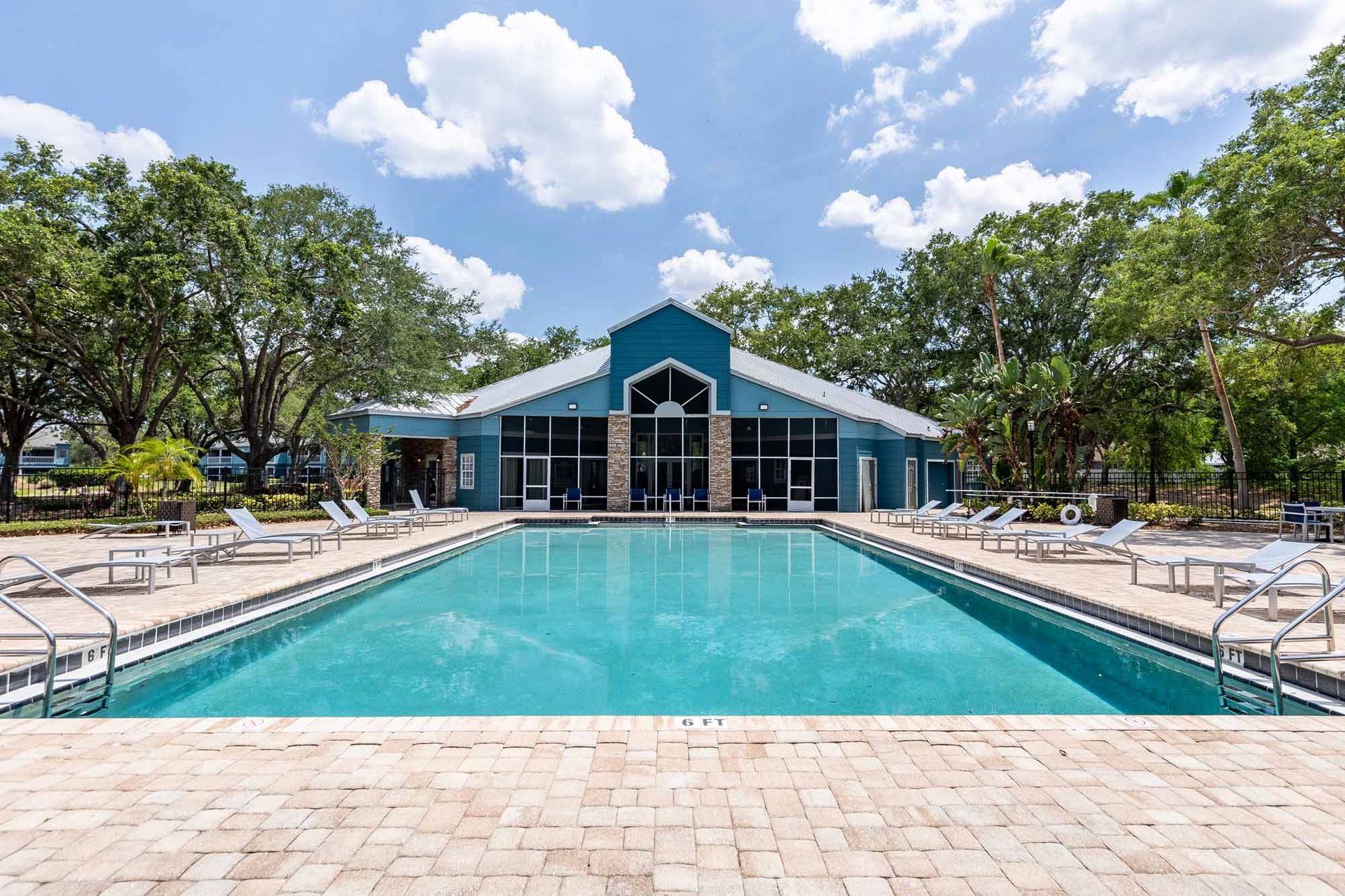 Pool with lounge chairs, facing a teal building under a blue sky with clouds.