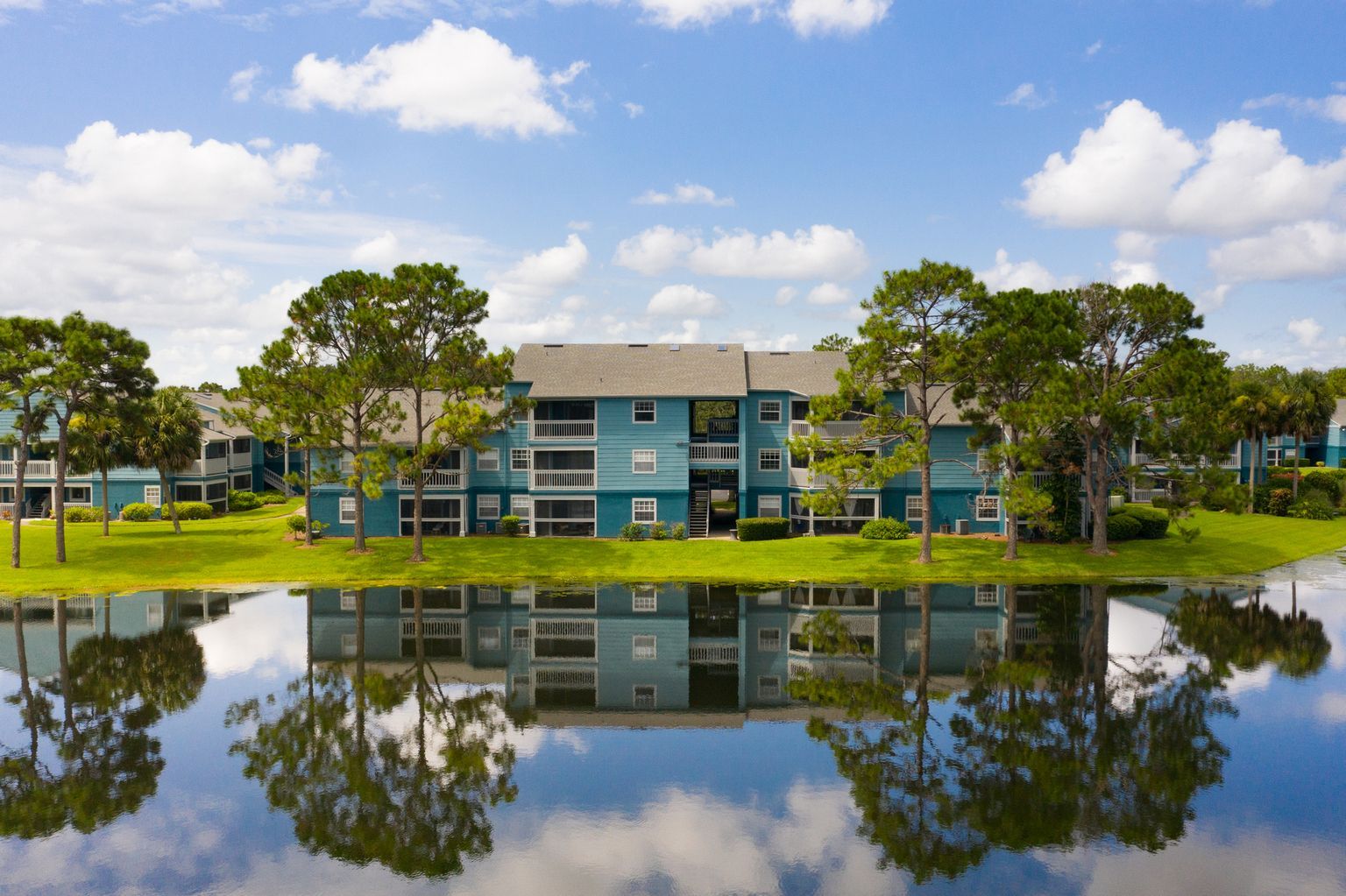 Blue apartment buildings reflected in calm water under a cloudy blue sky.
