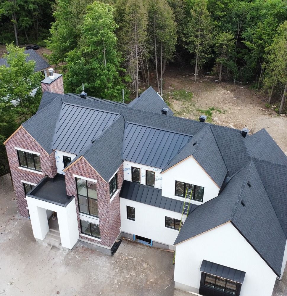 Aerial view of a large modern house with dark roof, white walls, and brick accents beside a wooded area.