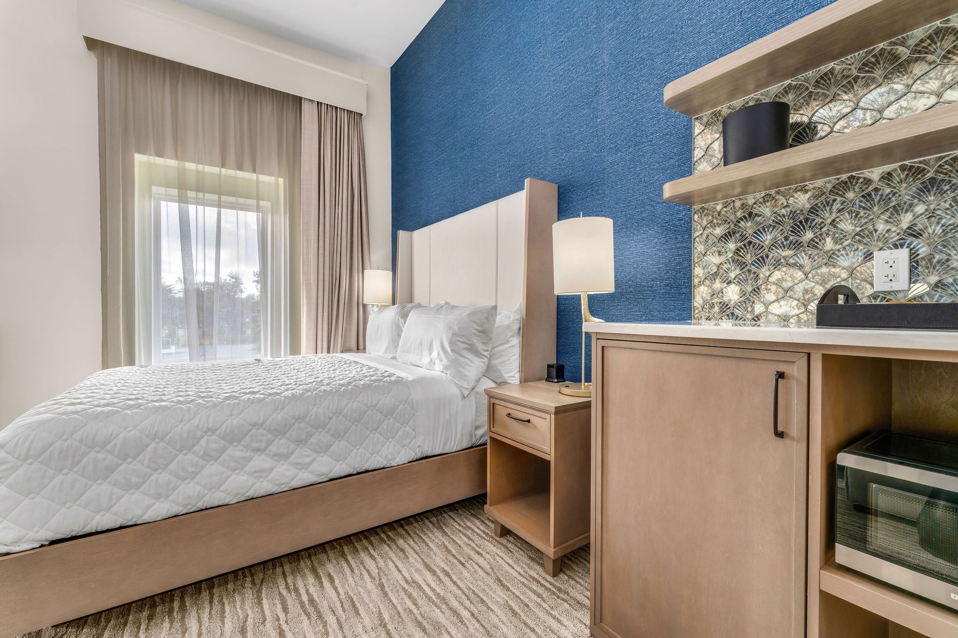 Queen size bed positioned against deep blue accent wall with nightstand & decorative lamp. Kitchenette to the right side with microwave, shelving and coastal background.
