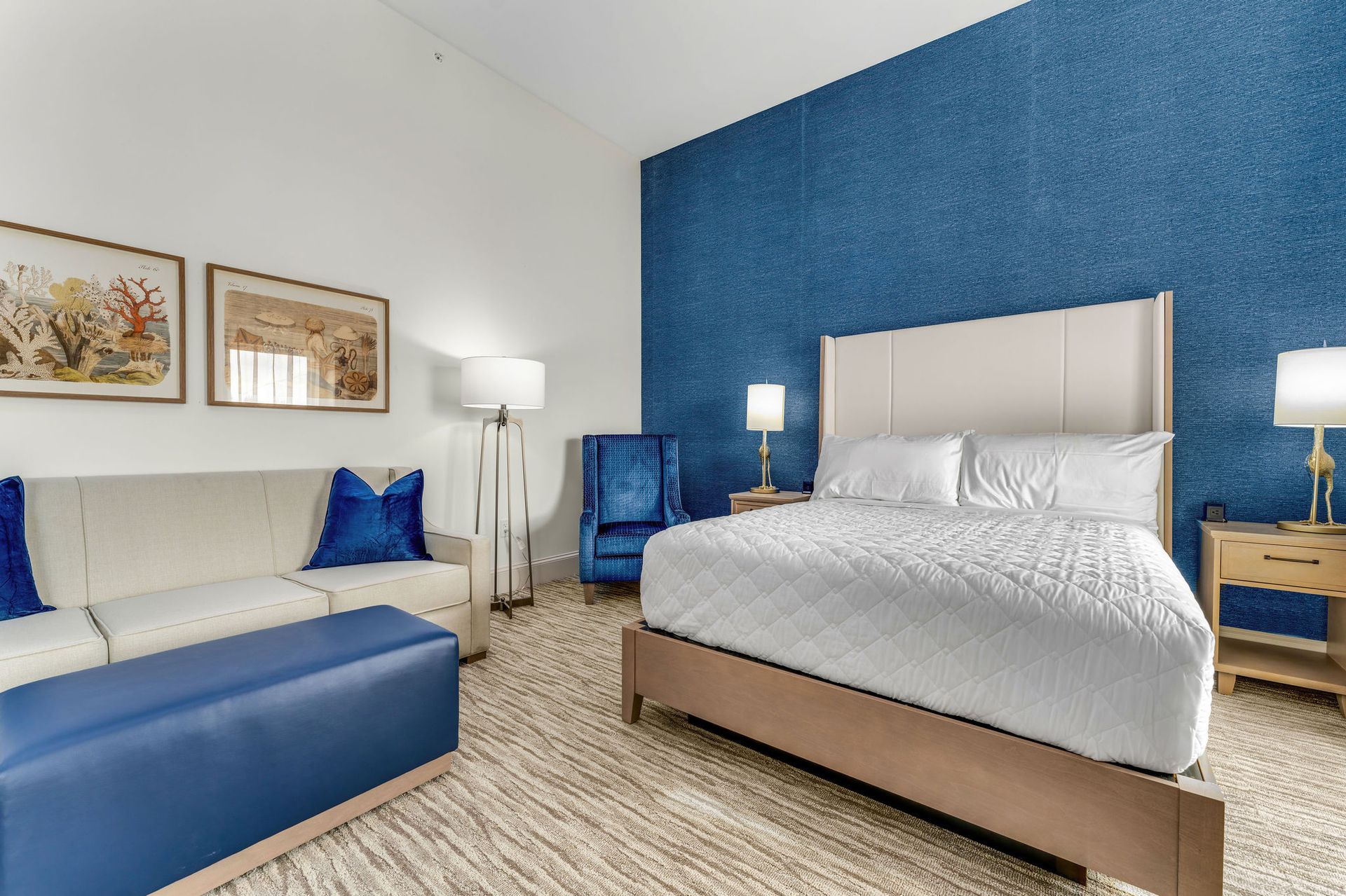 Queen size bed arranged in front of deep blue accent walls with nightstands & lamps on both sides of the bed. Floor lamp as well as light colored, full size couch with deep blue throw pillows, ottoman and plush accent chair.