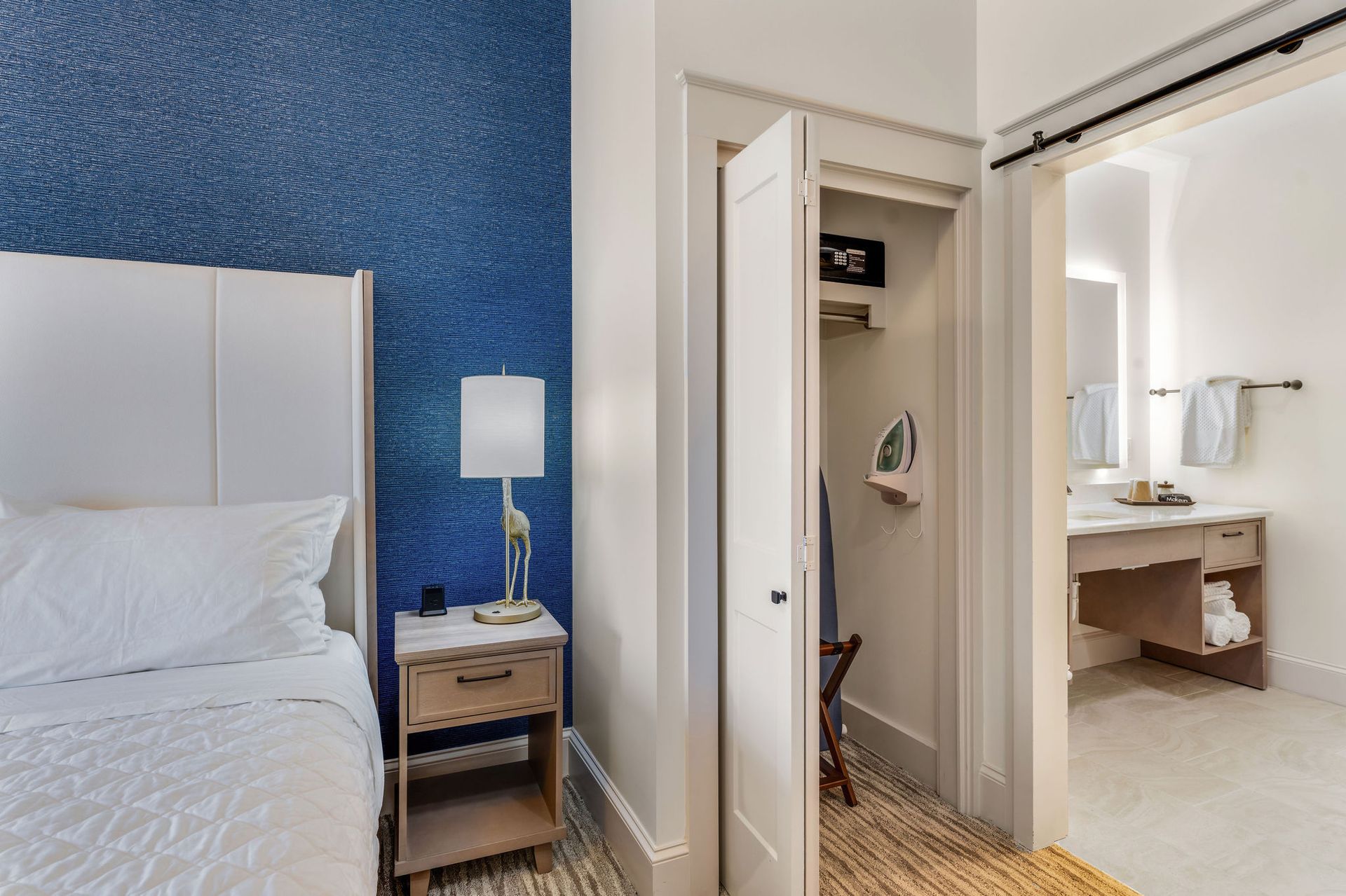 Nightstand with decorative lamp to the side of the bed. Closet to the right of the nightstand. The closet door is open showing a spacious closet with an iron, safe and a place to sit your luggage.