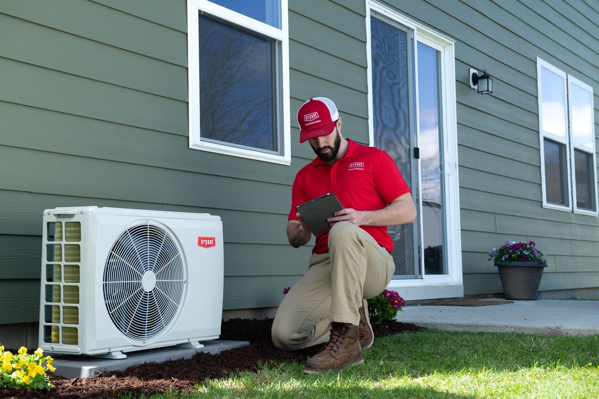HVAC technician kneeling by outdoor unit, inspecting with a tablet, near a green house.